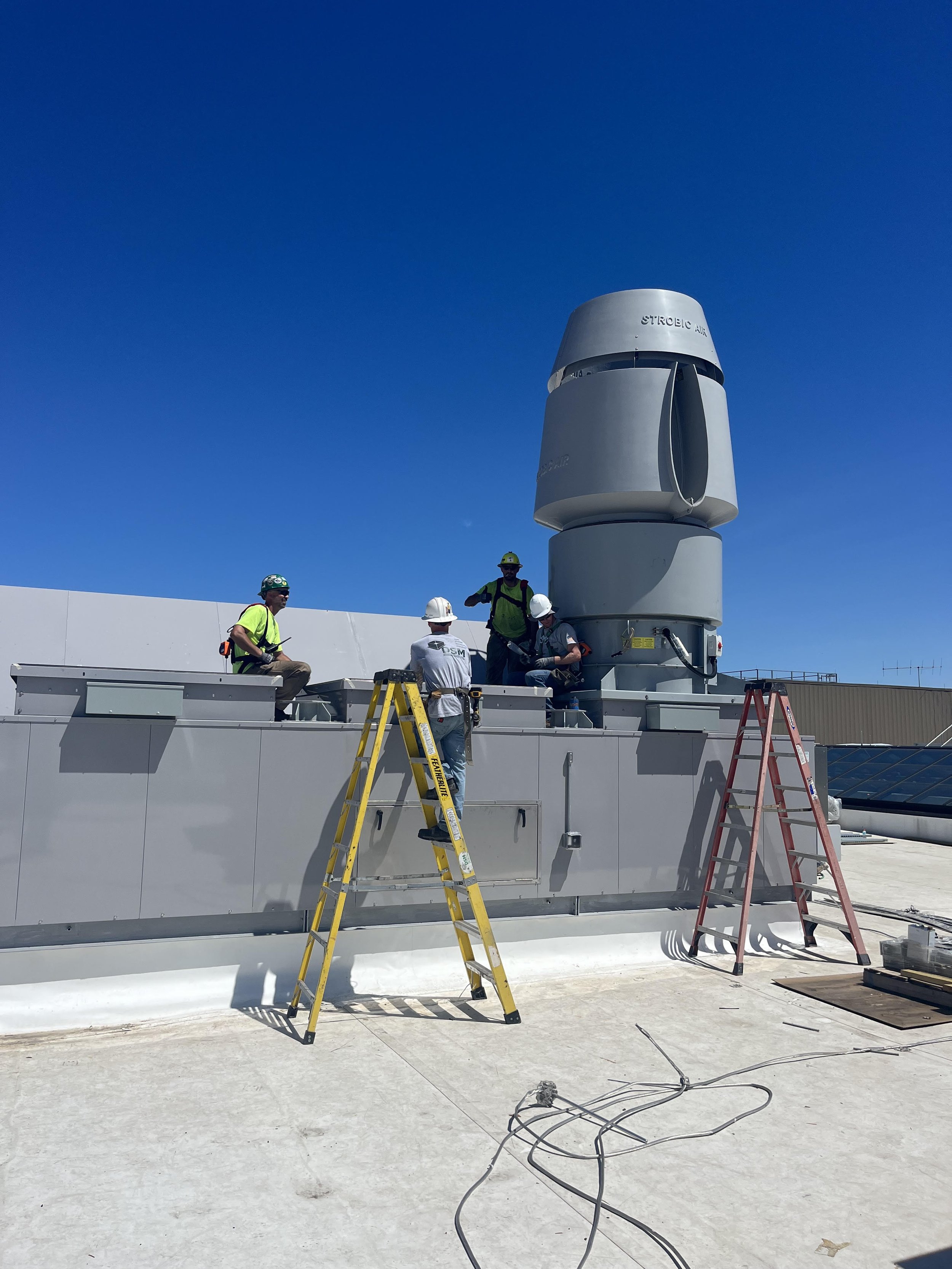 Workers on a rooftop installing or maintaining a large mechanical ventilation system under a clear blue sky, with some ladders and electrical cables on the ground.