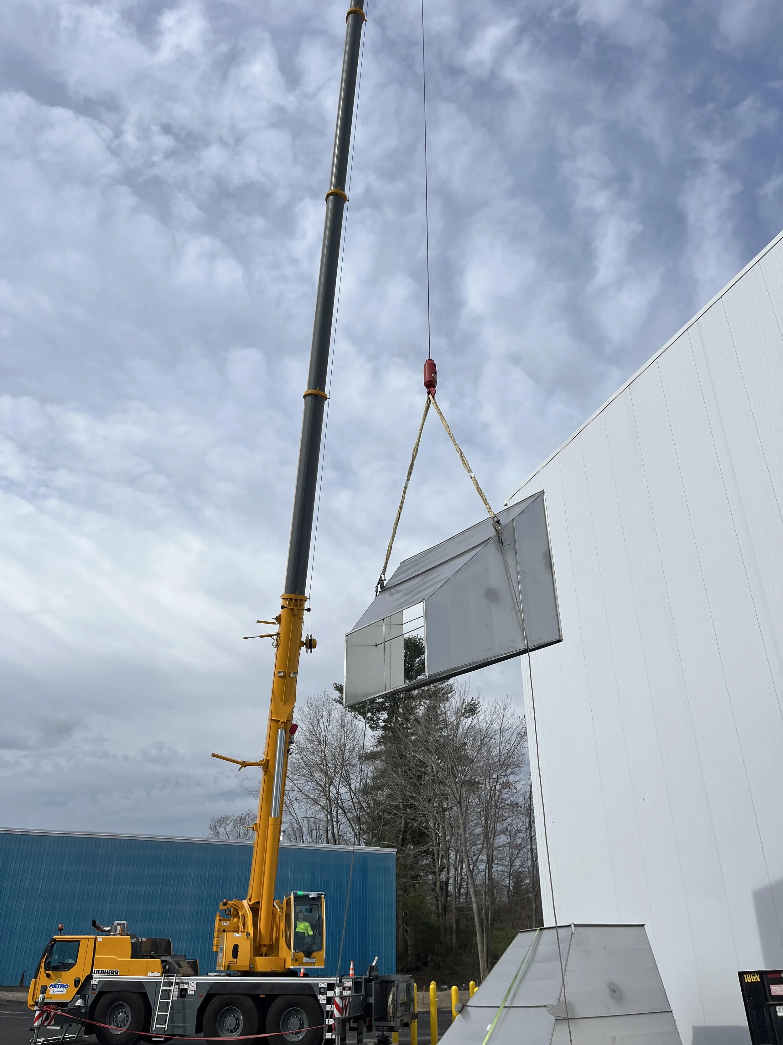 A large yellow crane lifting a long gray structure onto a building outside. The sky is cloudy and there are leafless trees in the background.
