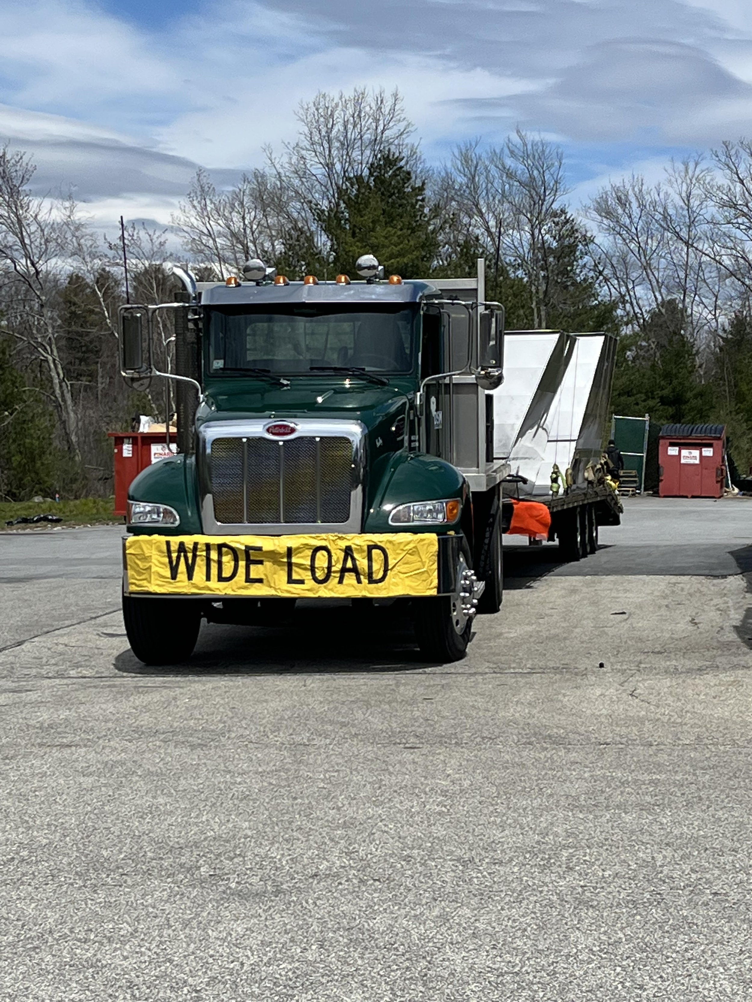 A large green truck with a yellow sign that reads 'WIDE LOAD' parked in front of a construction site. The background features trees, construction containers, and workers wearing safety gear.