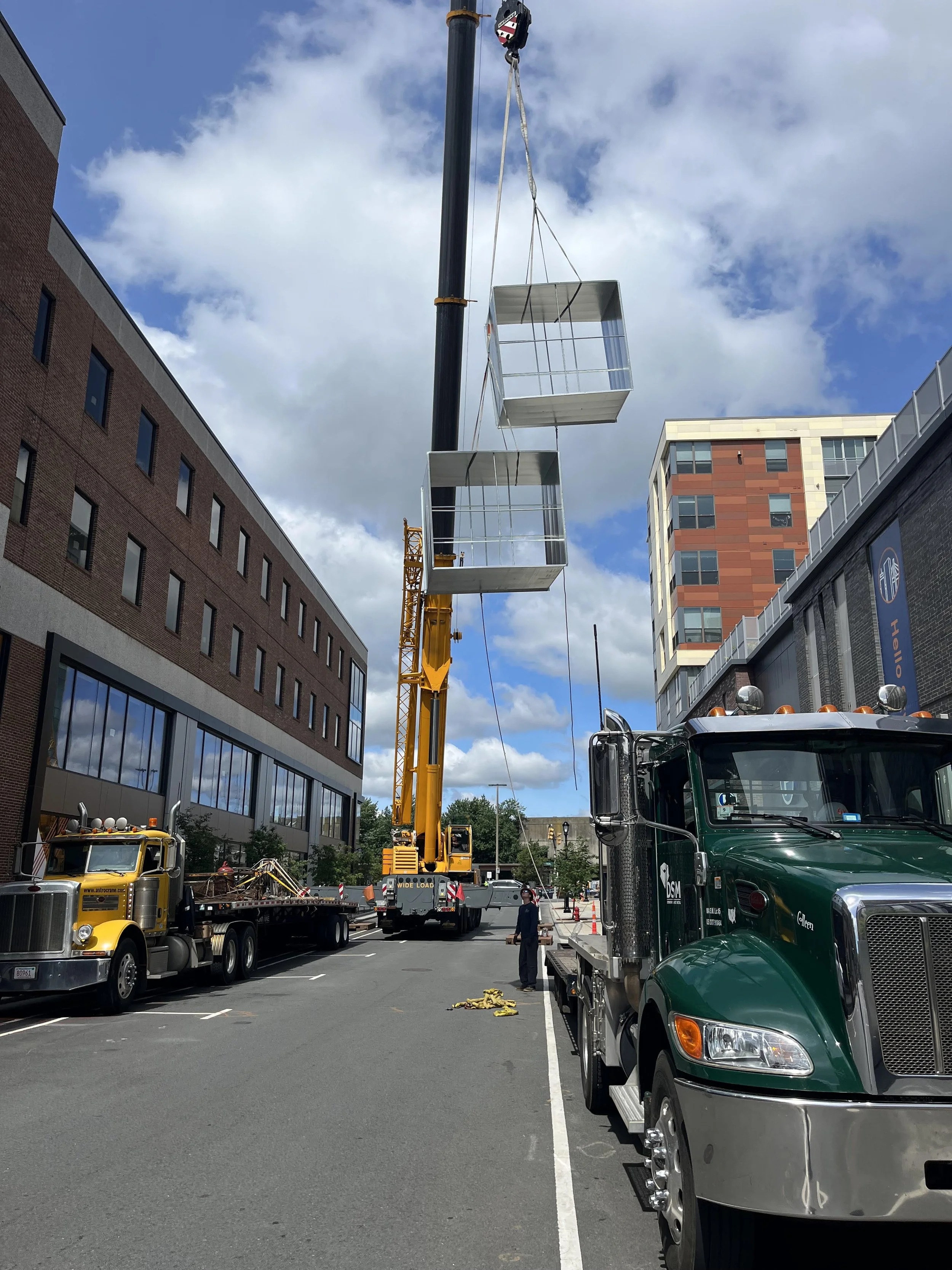Construction site on an urban street with a crane lifting a large glass structure, several trucks parked on the road, and modern buildings under a partly cloudy sky.