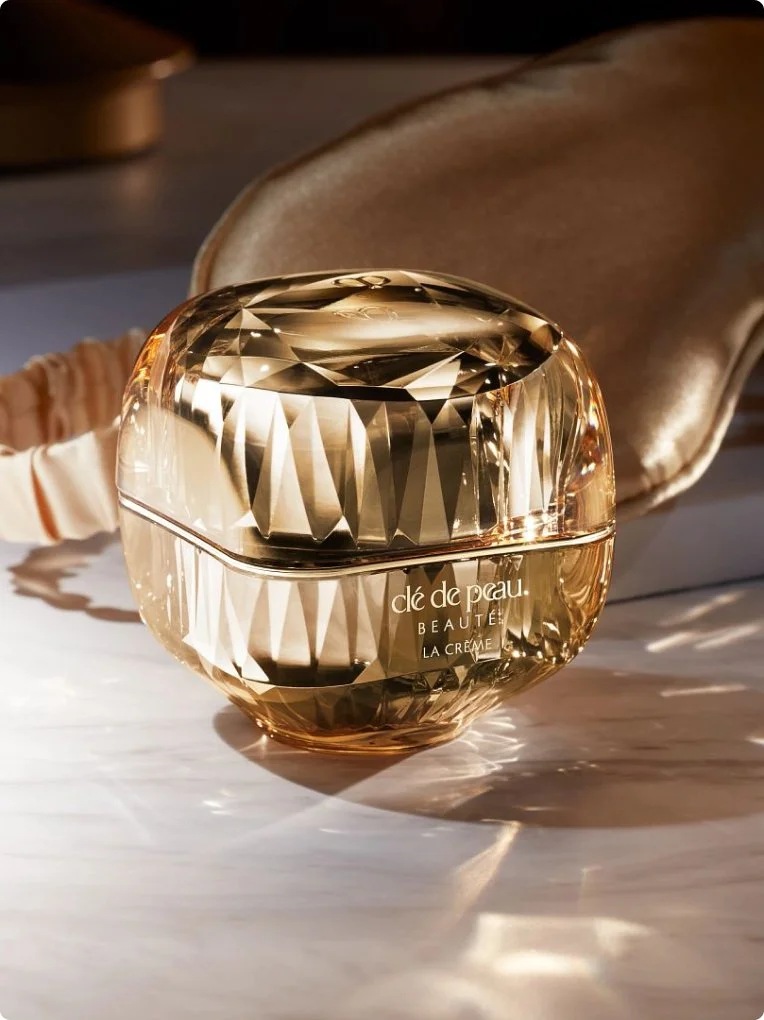 A luxurious glass jar of Clé de Peau Beauté La Crème cream on a reflective surface with a beige velvet pouch in the background.