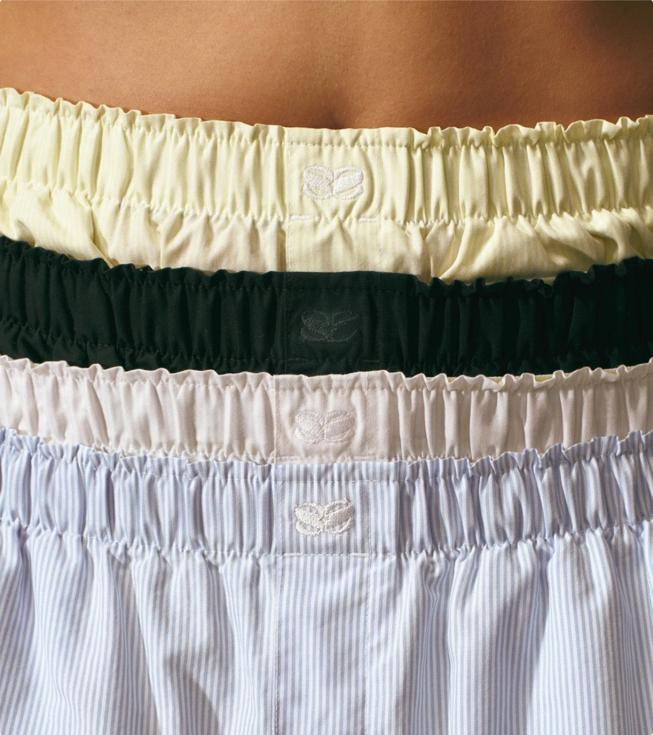 A close-up of four different pairs of men's boxers with elastic waistbands, stacked vertically. The top pair is light yellow, the second is black, the third is light beige, and the bottom is light blue with white stripes. Each waistband has a small embroidered lotus flower logo in white.