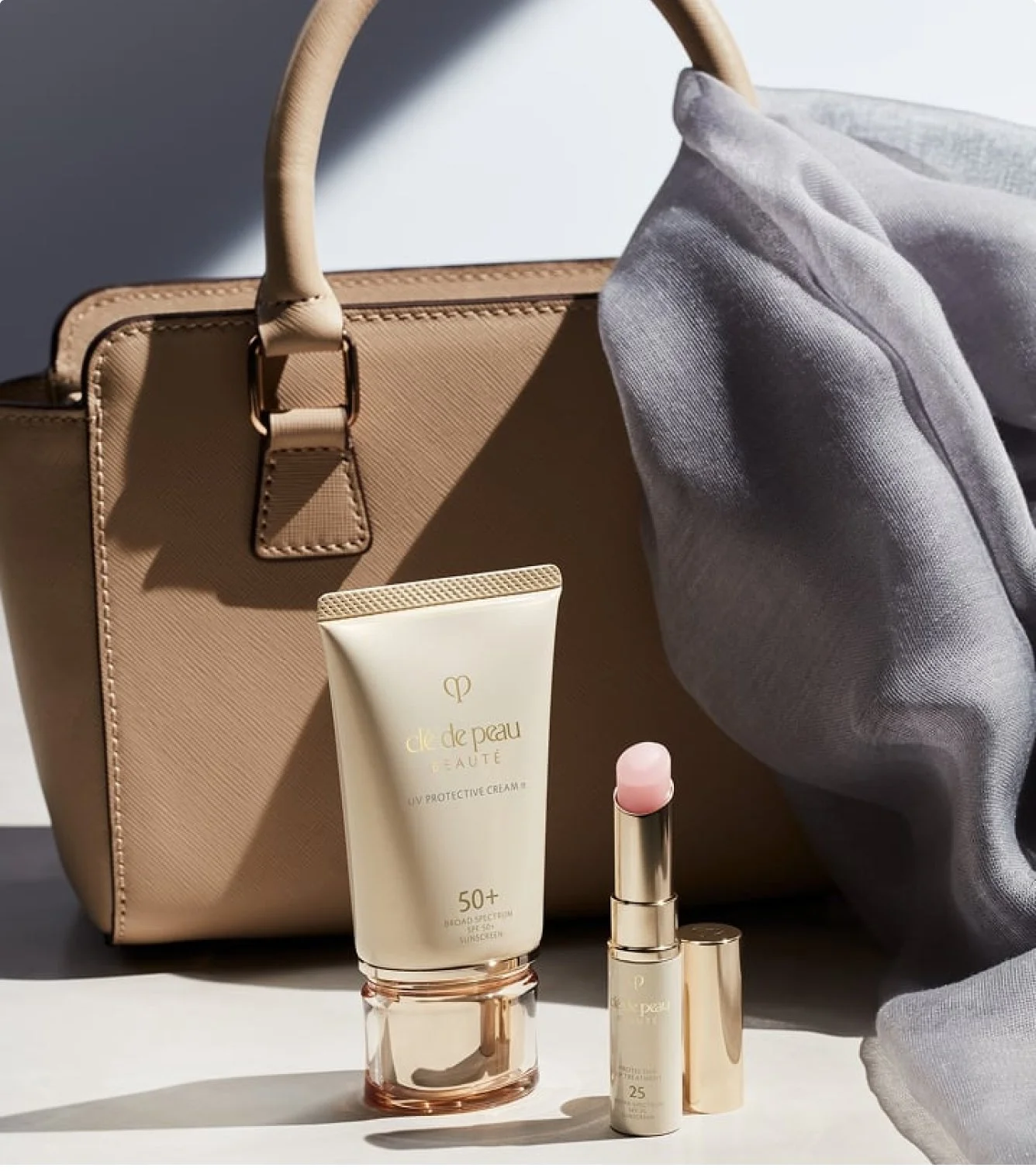 A beige handbag with a textured surface and brown hardware, a tube of Clé de Peau Beaute UV protective cream, a pink lipstick, and other skincare or makeup products are arranged in front of a gray fabric item, with a gray cloth partially draped over the handbag.