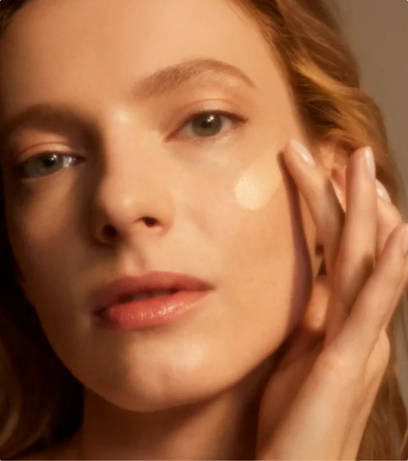 Close-up of a woman with light skin, blonde hair, and hazel eyes touching her face gently.