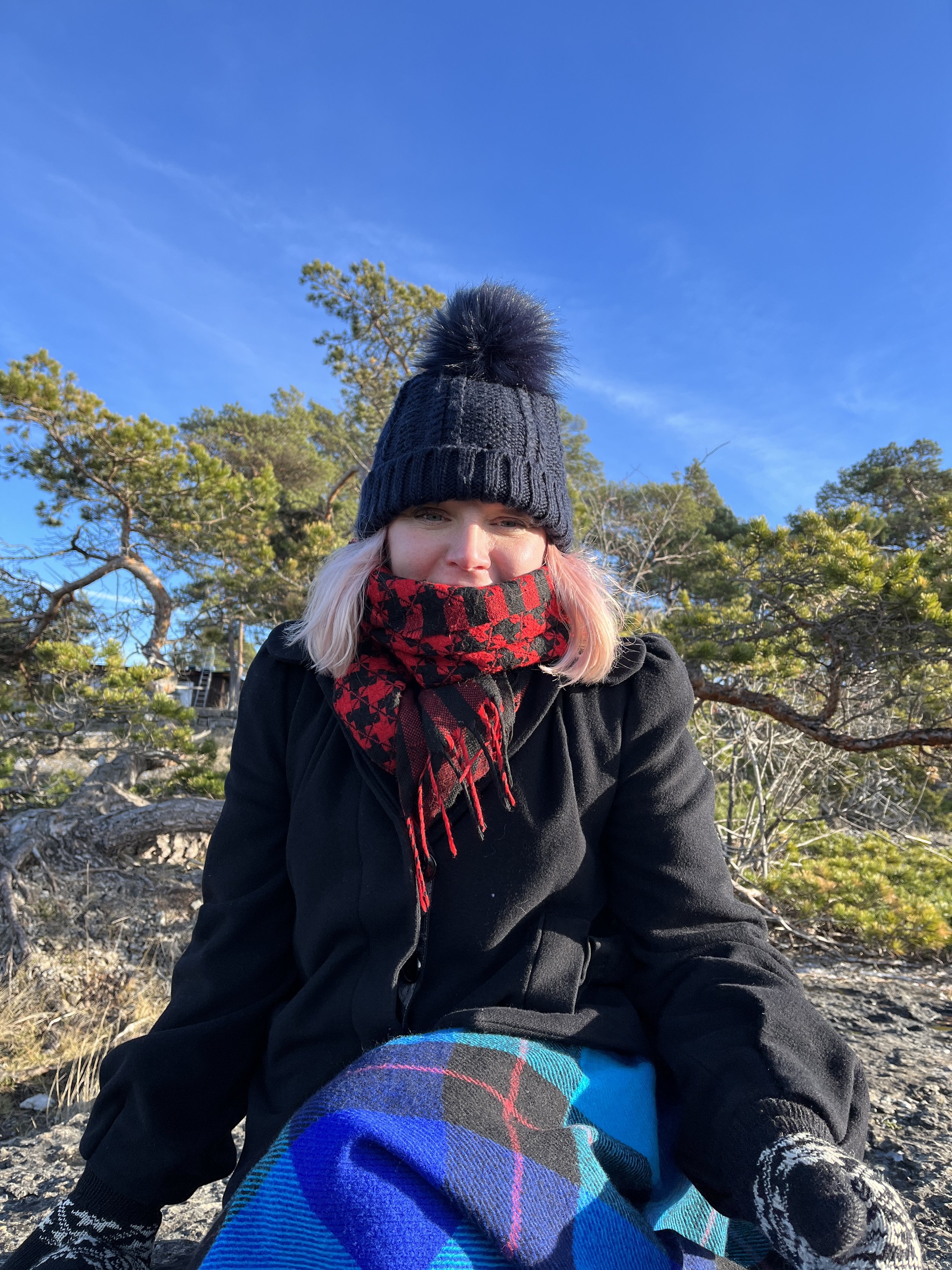 A person with pink hair wearing a black beanie with a pom-pom, a red and black scarf, and a black jacket sitting outdoors among trees under a blue sky.