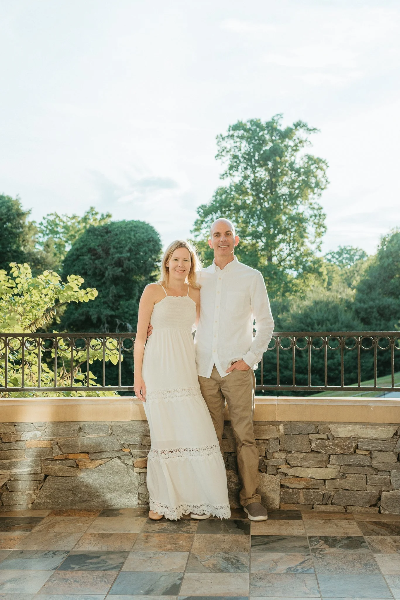 A couple posing outdoors on a balcony with greenery and trees in the background, colorful tiled floor, stone wall, and railing, during daytime with bright sky.