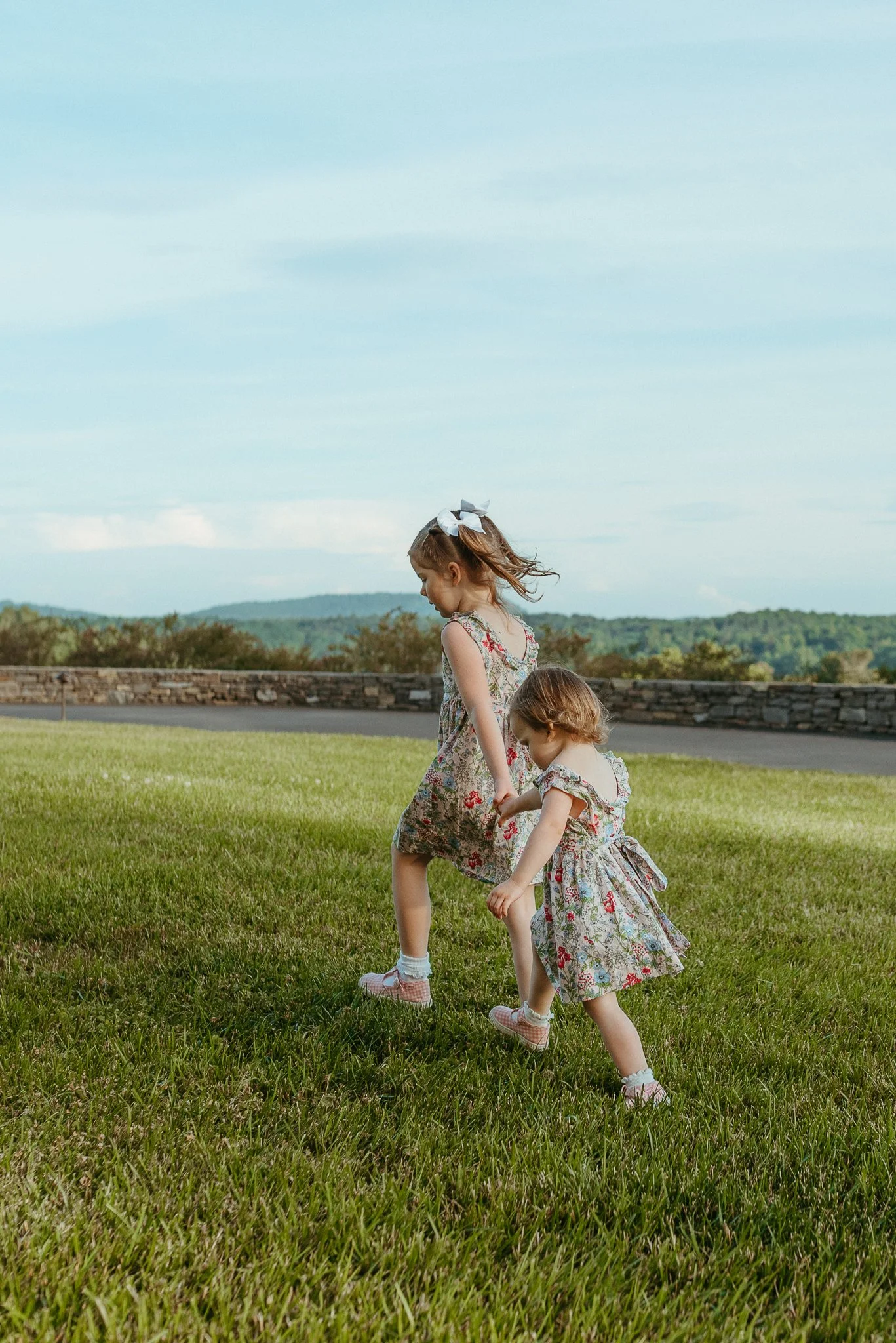 Two young girls holding hands and playing in a grassy field on a clear day. Both girls are wearing floral dresses and sneakers, with one girl having a large white bow in her hair.