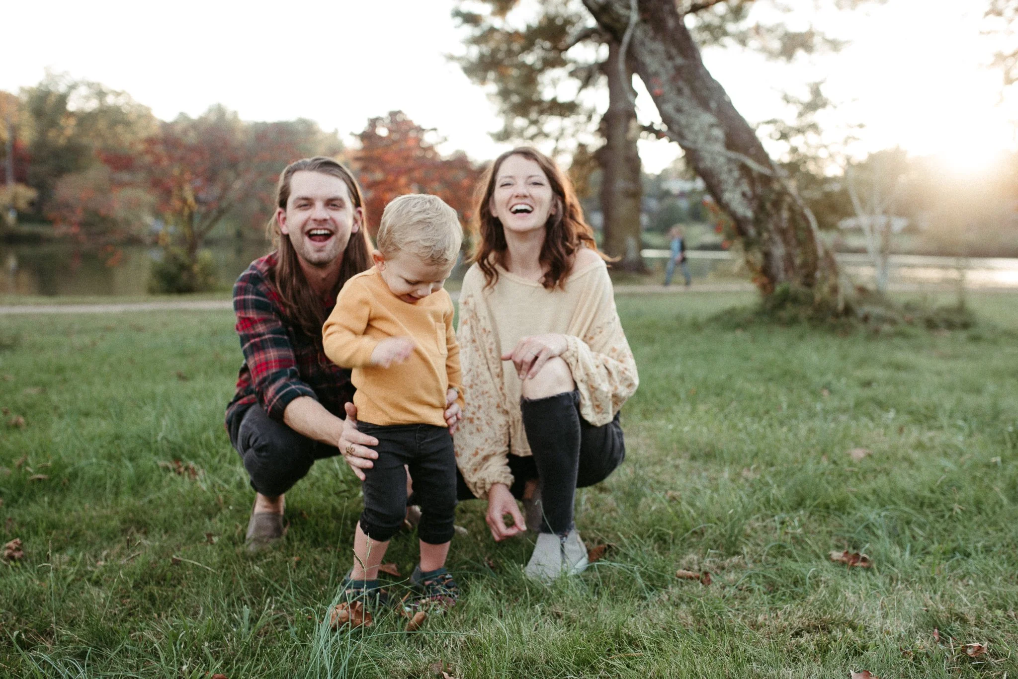 A happy family of three—a man, woman, and young boy—are outdoors in a park during autumn, smiling and playing on the grass with trees and a pond in the background at sunset.