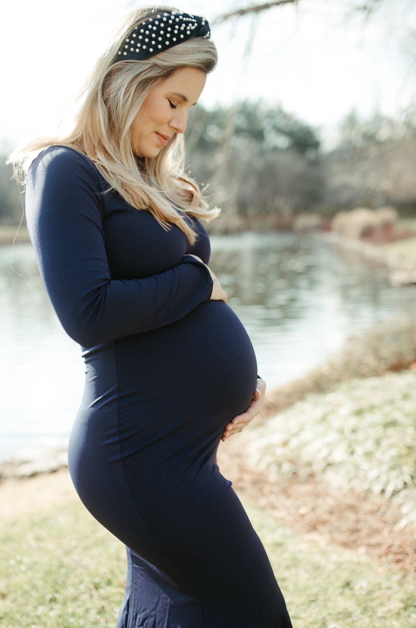 Pregnant woman with blonde hair wearing a dark blue dress and a black headband with white polka dots, standing outdoors near a body of water and trees, with hands on her belly and a peaceful expression.