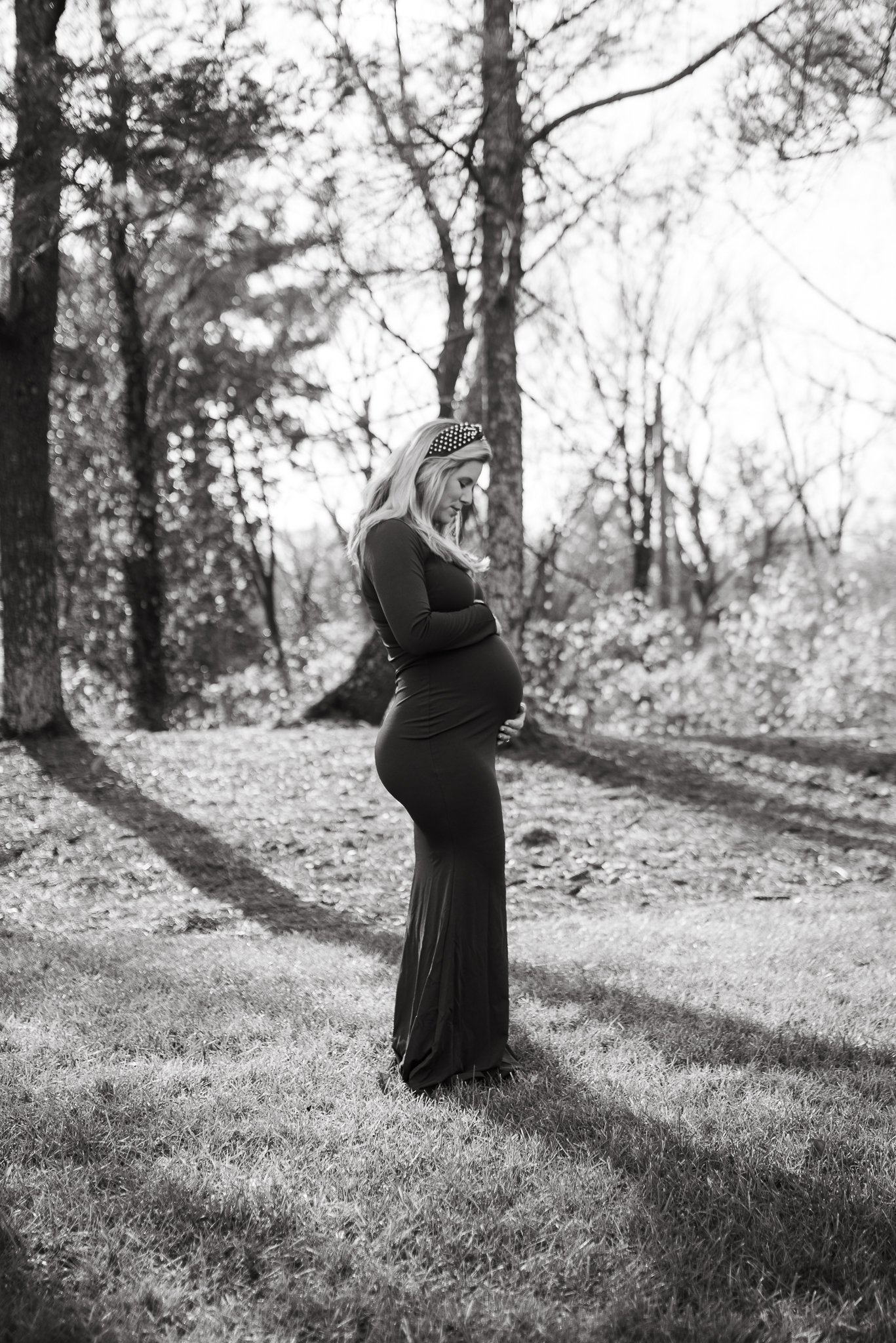 A pregnant woman standing outdoors on grass, gently holding her baby bump, with trees in the background, black and white photograph.