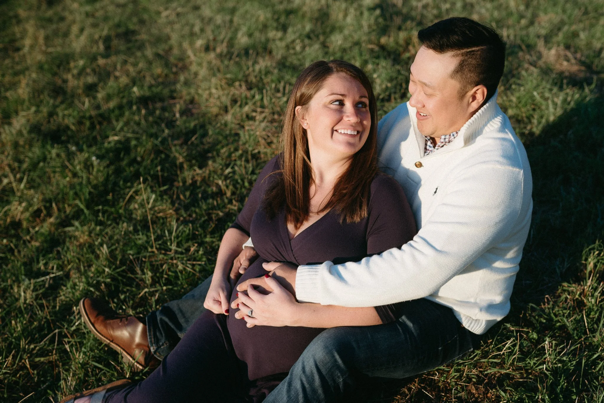 A couple sitting on grass, smiling and looking into each other's eyes, with their arms around each other outdoors.