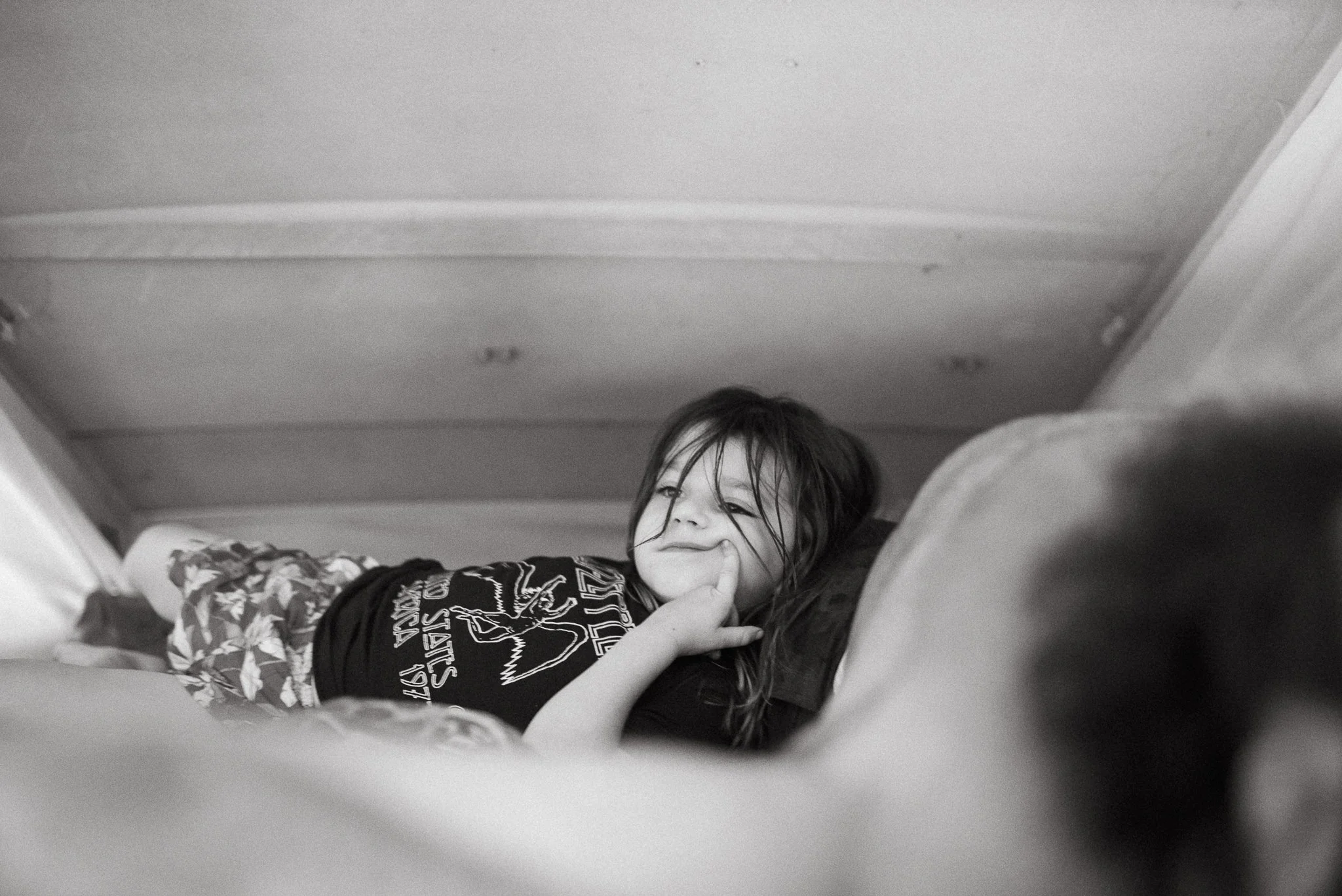 A young girl with long hair lying on her stomach, smiling and resting her head on her hand, in a room with a plain ceiling and partial view of her clothes and arm.