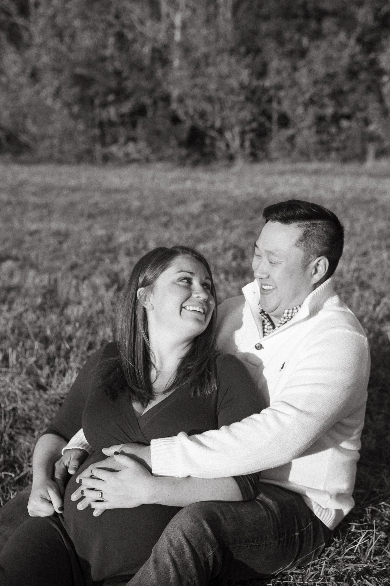 A happy couple sitting on grass outdoors, looking into each other's eyes and smiling. The woman has long hair and is wearing a dark top, while the man has short hair and is wearing a light sweater. They seem to be enjoying a joyful moment together.