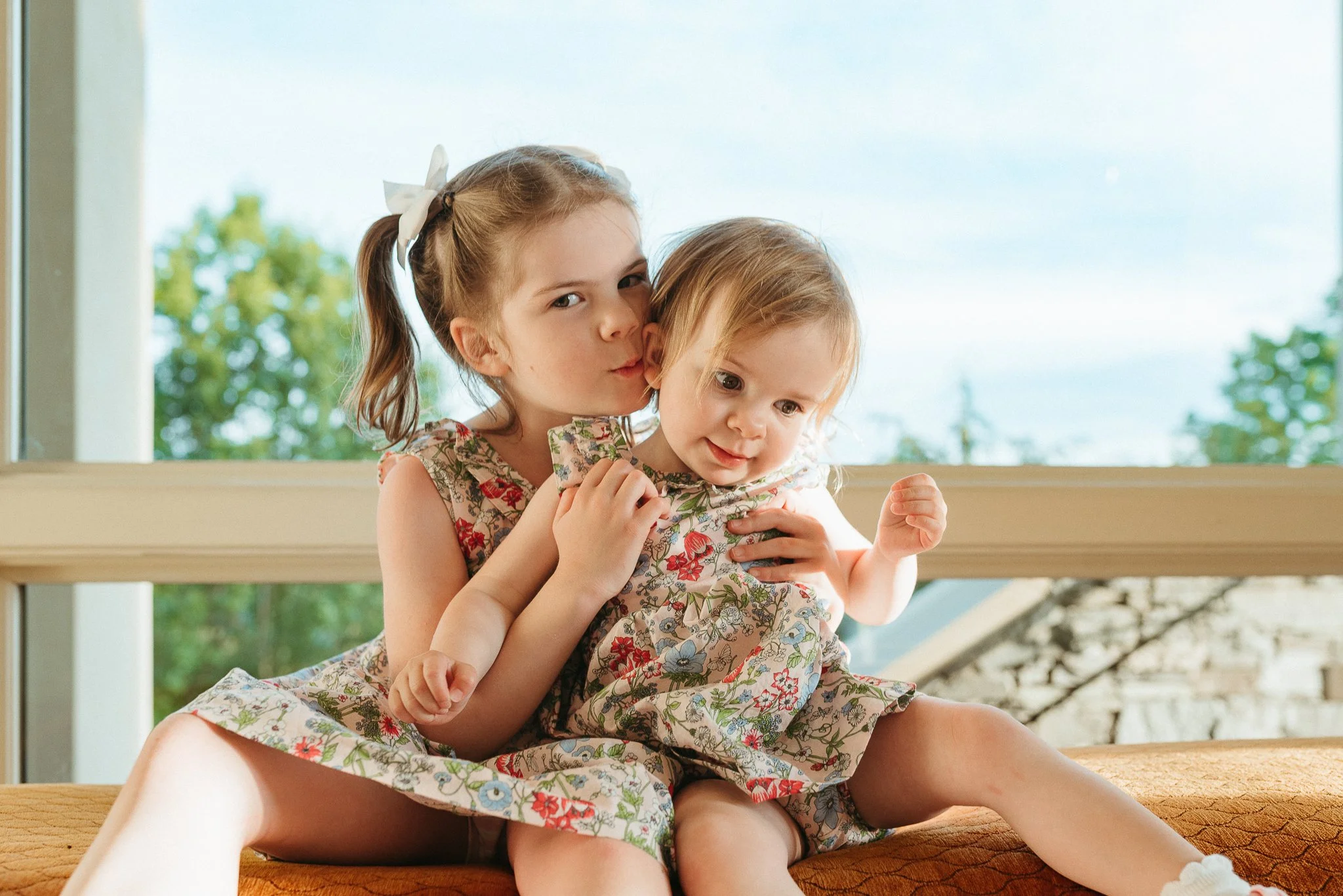 Two young girls, one older and one younger, sitting together on a wooden surface near a large window with greenery outside. They are wearing floral dresses and are close to each other, with the older girl holding the younger girl and giving her a gen
