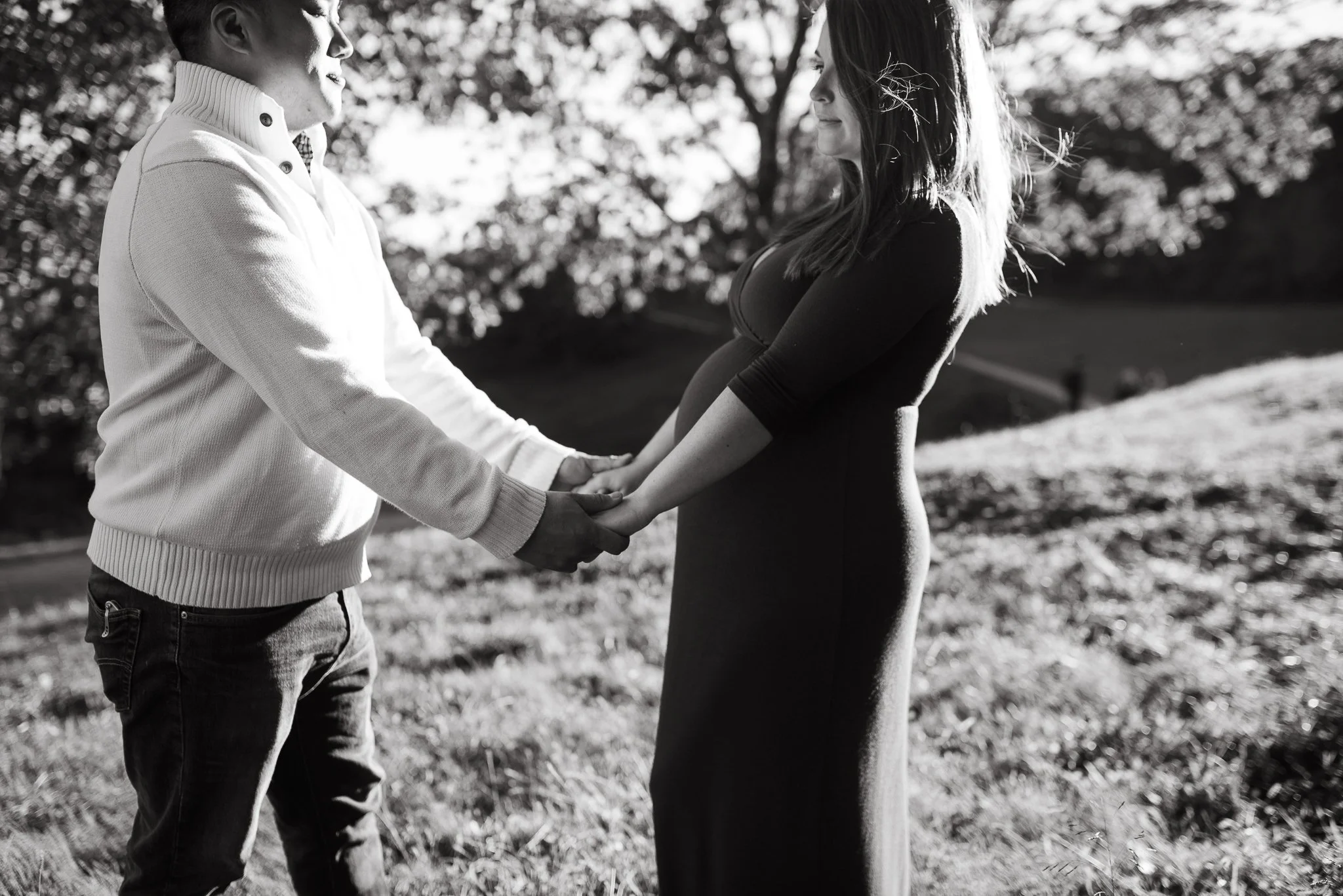An expectant couple standing outdoors holding hands, with the woman’s baby bump visible. The woman has long hair and is wearing a fitted dress, while the man is wearing a sweater and jeans. The background features trees with sunlight filtering throug