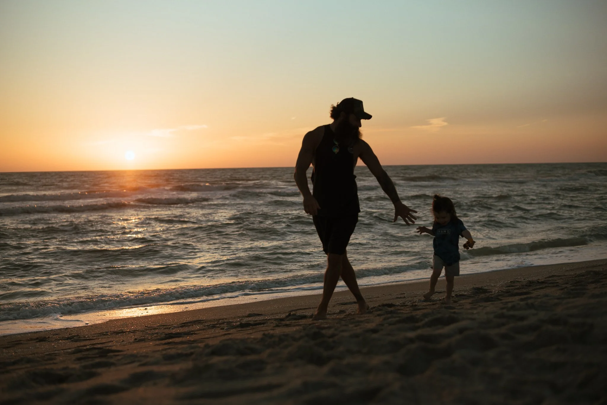 A man and a young girl walking on the beach at sunset with the ocean and sky in the background.