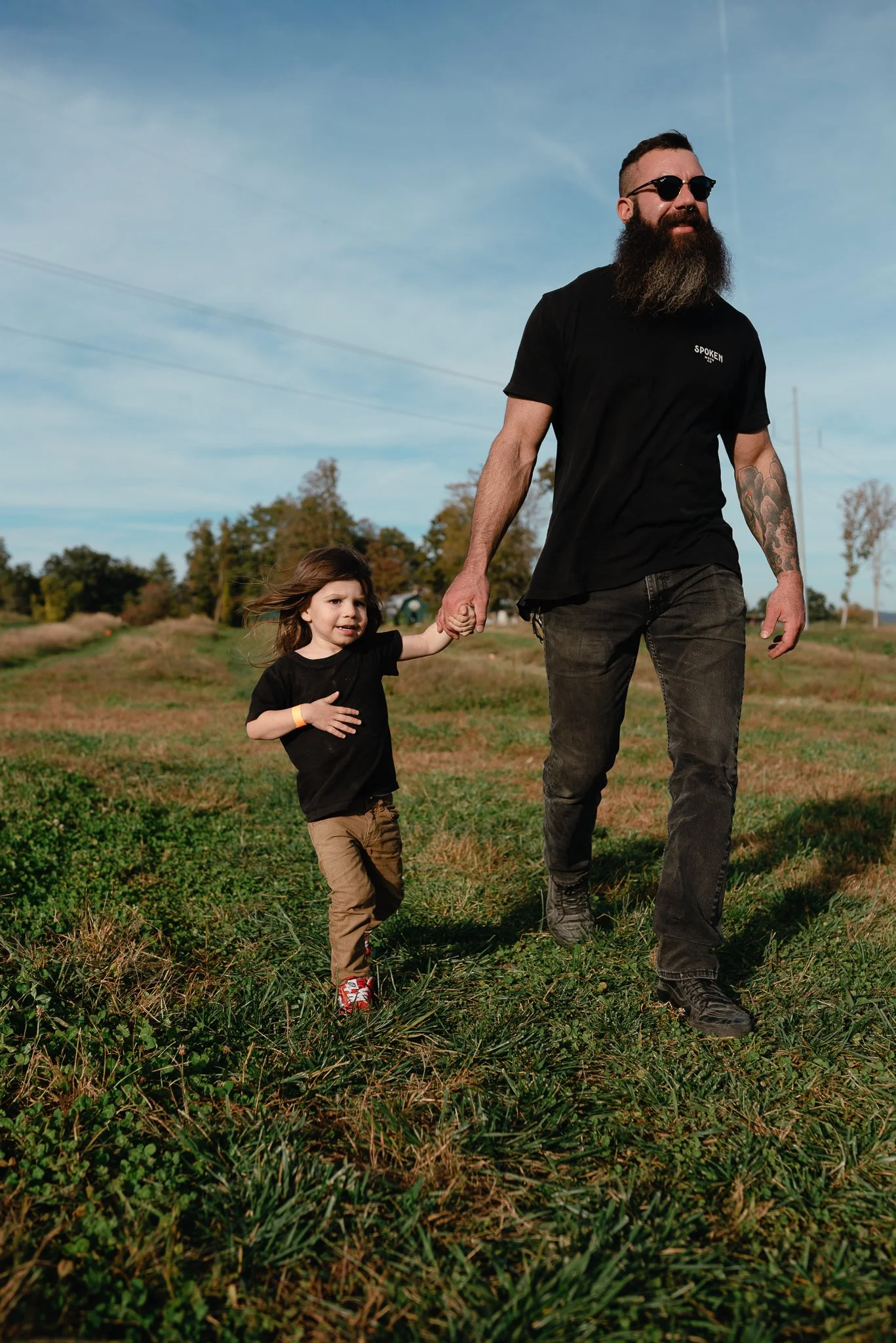 A man with sunglasses and a beard holding the hand of a young girl walking through a grassy field with trees and a clear blue sky in the background.