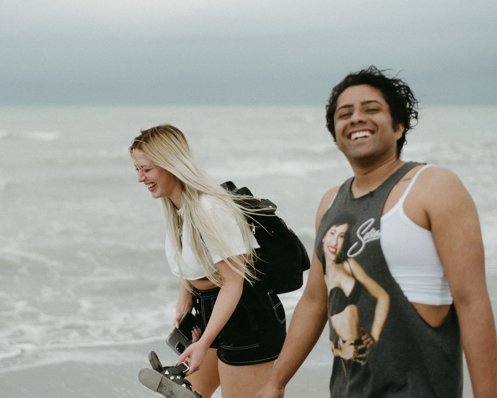Two young women and a young man walking along the beach, smiling, with the ocean and cloudy sky in the background.