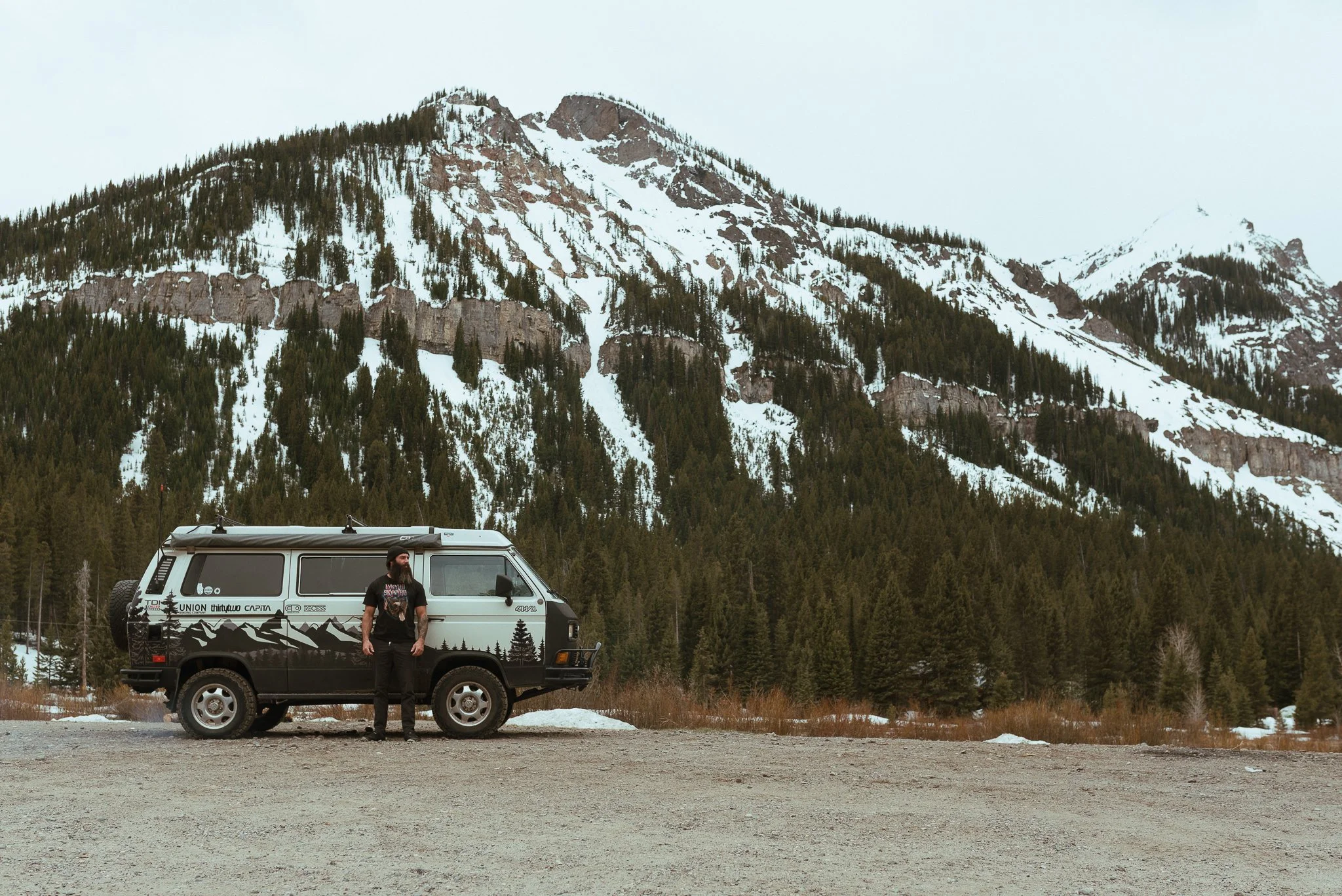 A man standing beside a camper van in a mountainous area with snow-covered peaks and dense pine trees.