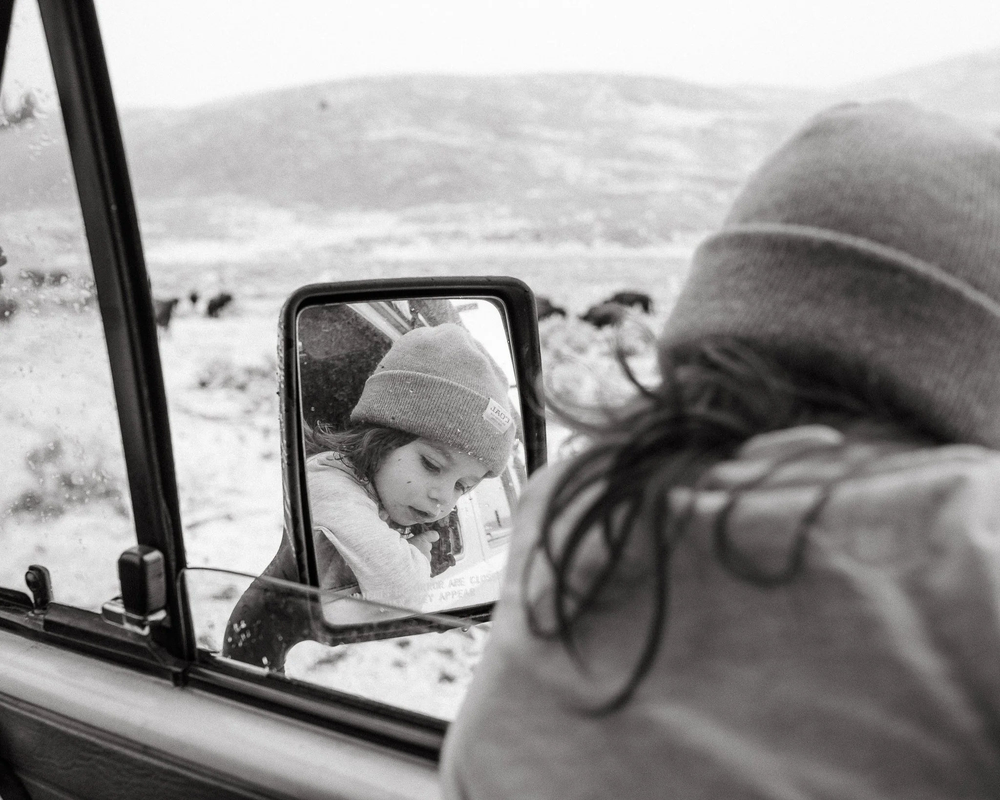 A person in a beanie and jacket looking at a snow-covered landscape through a mirror attached to snowmobile or ATV, with snow and mountain in the background.