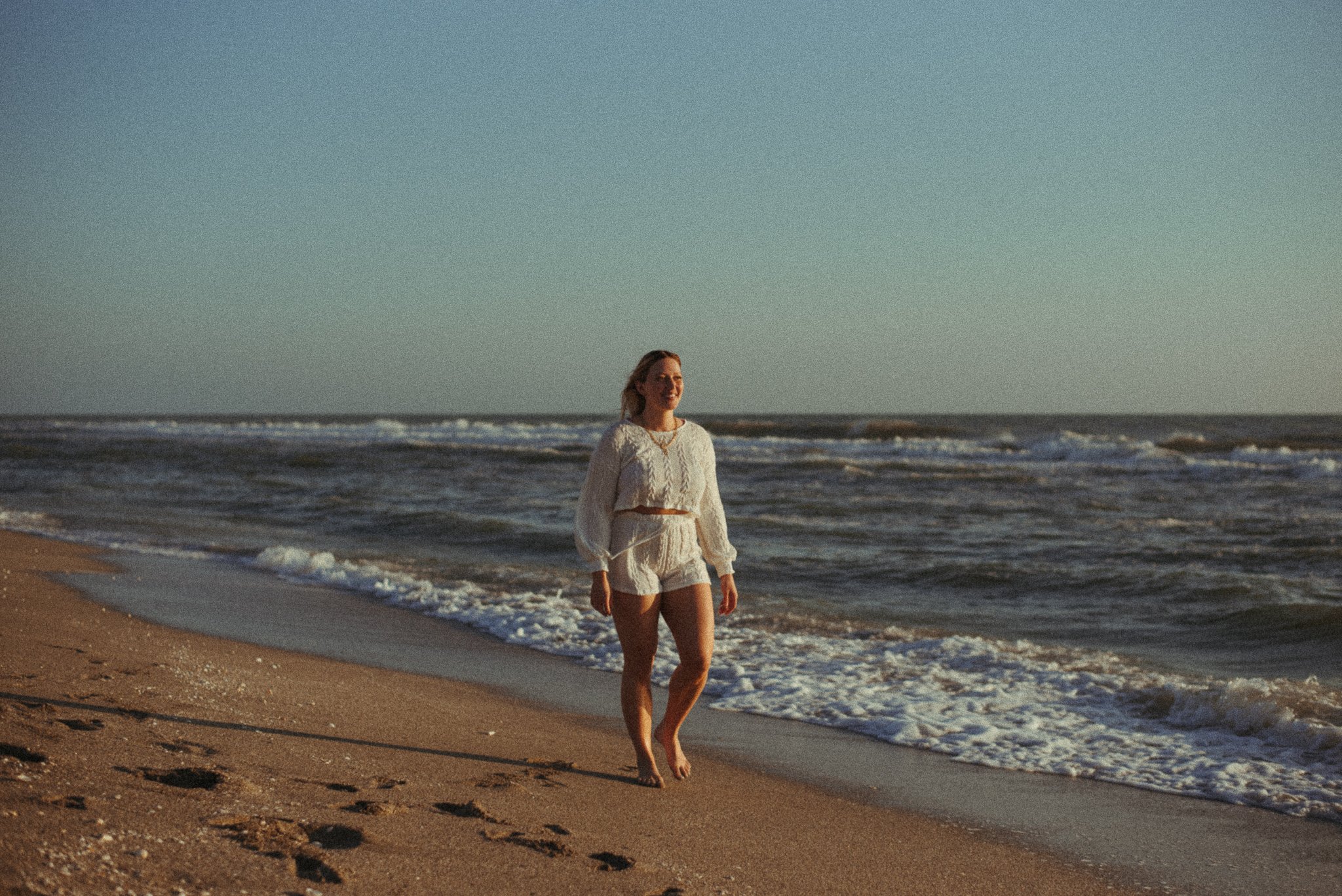 A woman wearing a white dress walking barefoot on the beach with waves in the background during sunset.