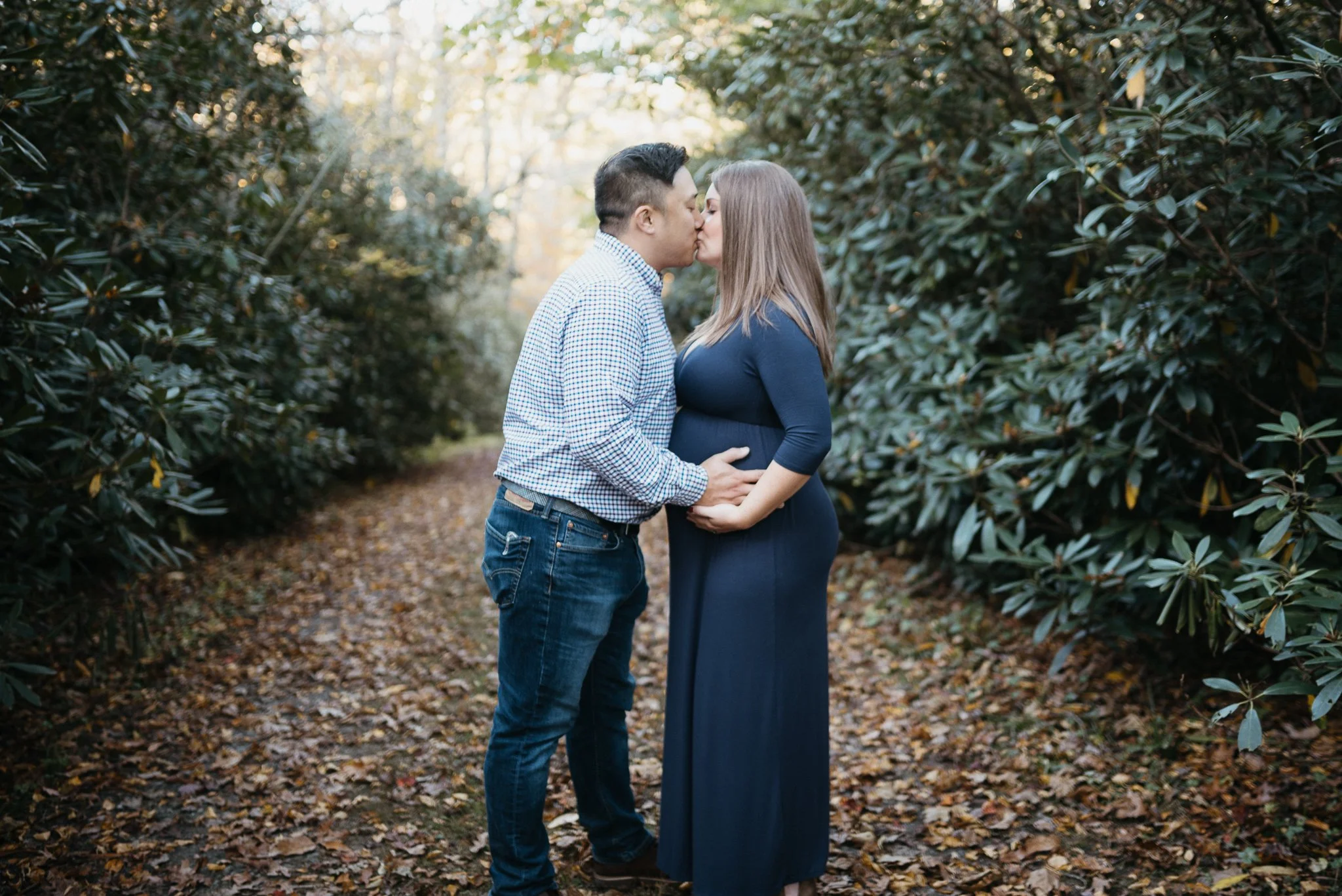 A couple kisses in a wooded outdoor setting, with fallen leaves on the ground and greenery on both sides.