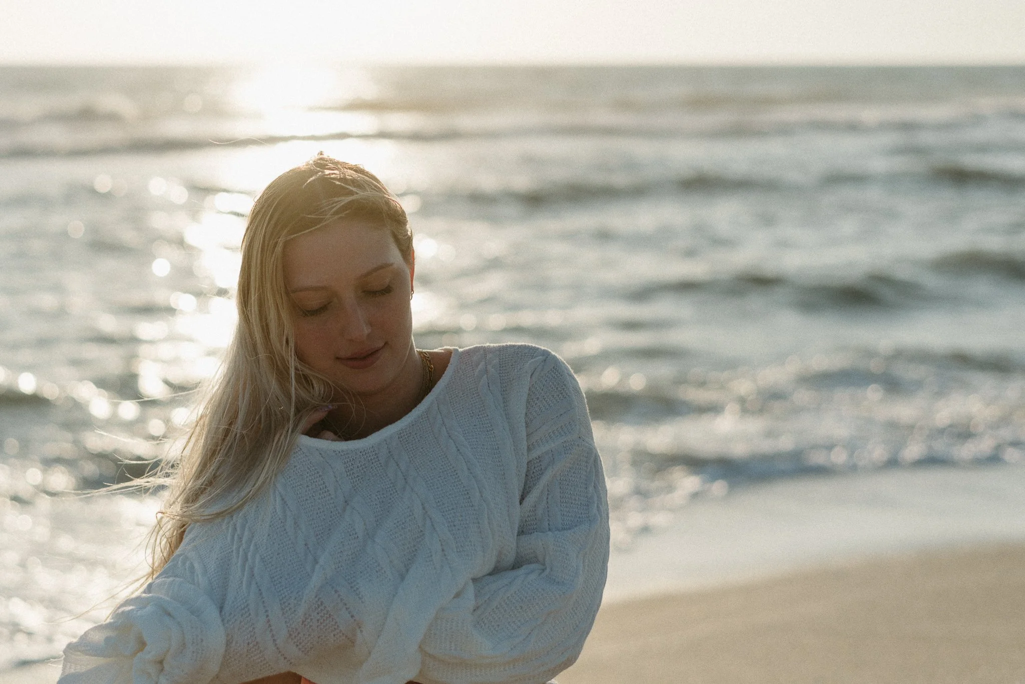 A woman with long blonde hair standing on a beach with the ocean in the background, wearing a white sweater.