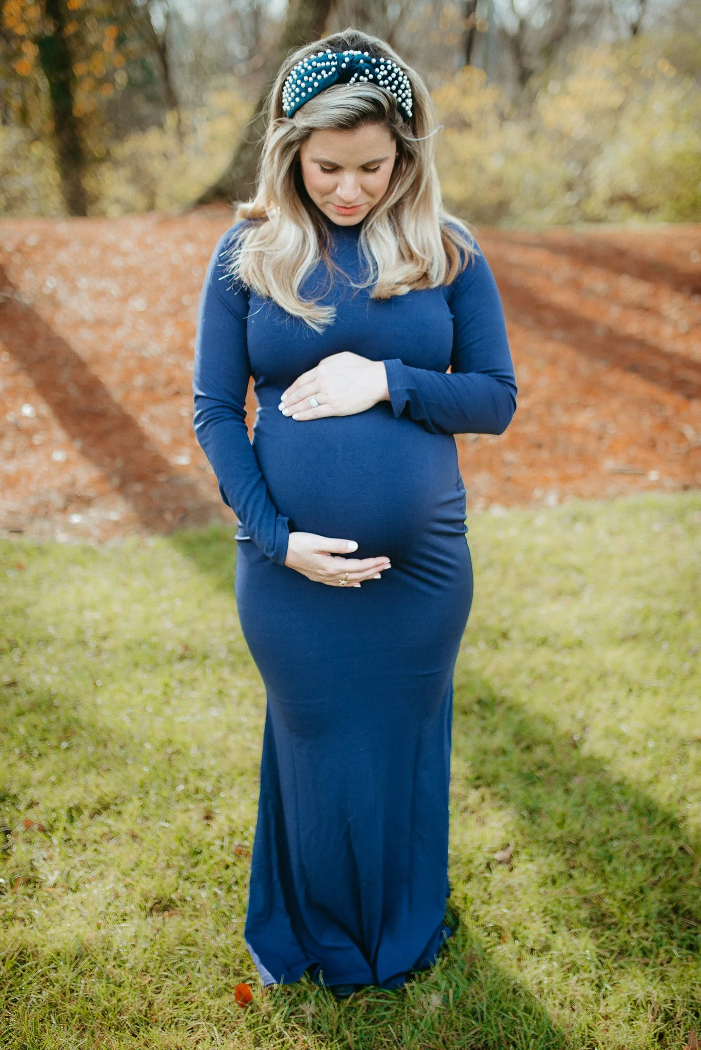 A pregnant woman in a long blue dress is standing outdoors in a park, gently touching her belly with both hands. She is wearing a headband with pearls and has blonde hair. The background features trees with fall foliage and a grass-covered ground.