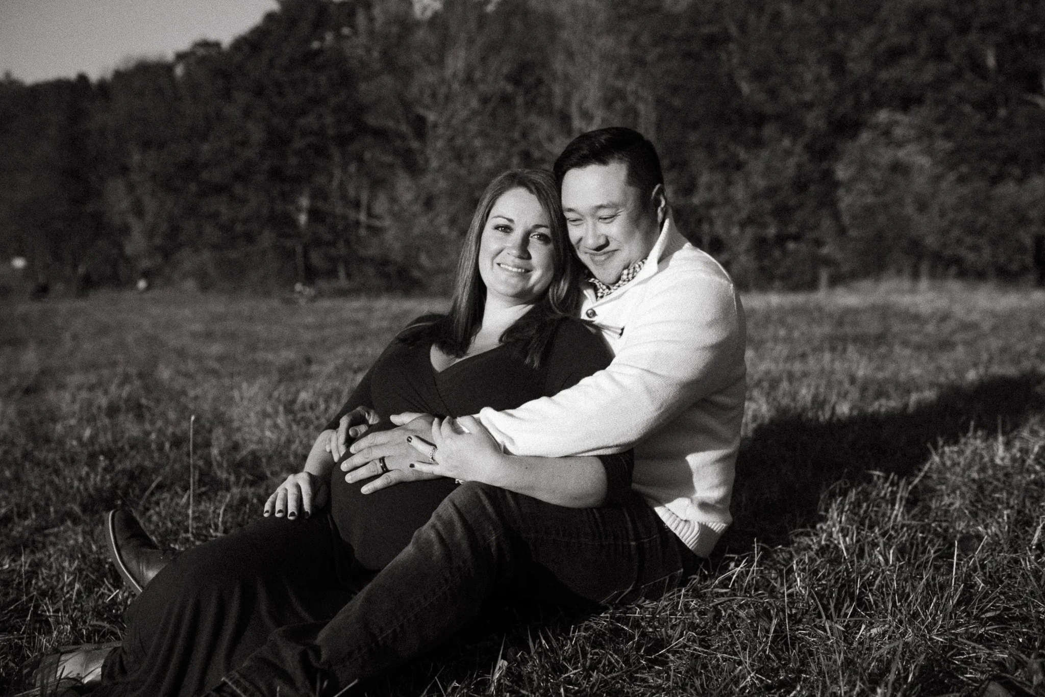 A black and white photo of a smiling woman and a man sitting on grass outdoors. The woman is leaning into the man, and they are embracing each other, with trees in the background.