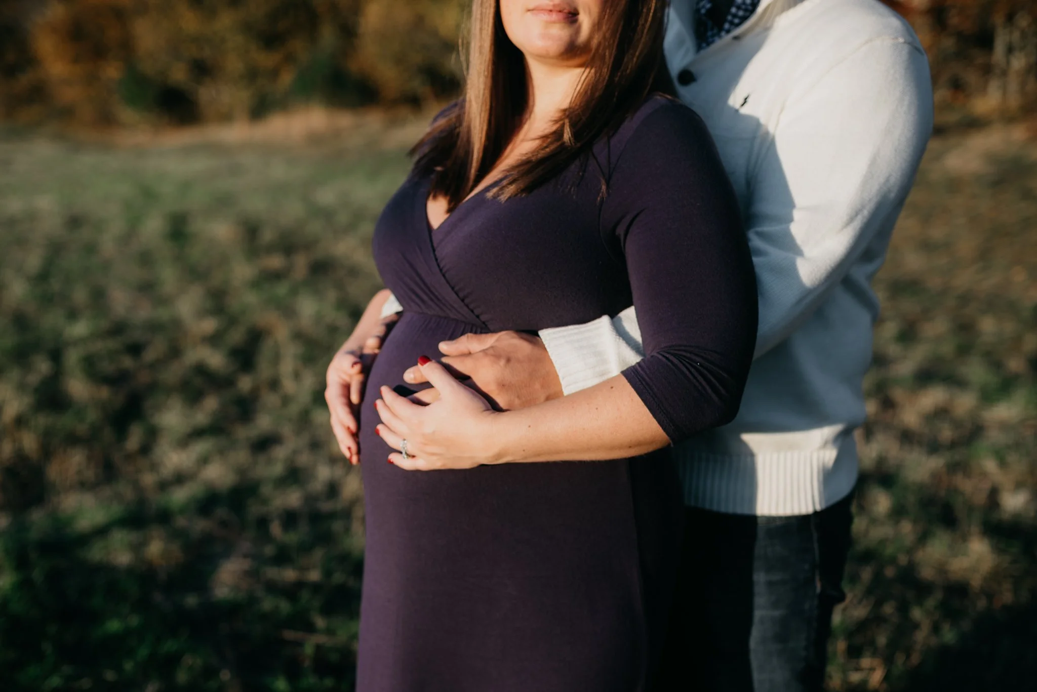 A couple standing outdoors, with the woman showing her pregnant belly and both holding her stomach together.