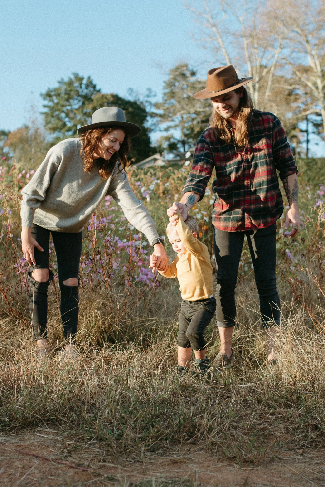 A family of four, including two women, one man, and a young boy, enjoying a walk through a field with tall grass and wildflowers on a sunny day, with trees and a house visible in the background.