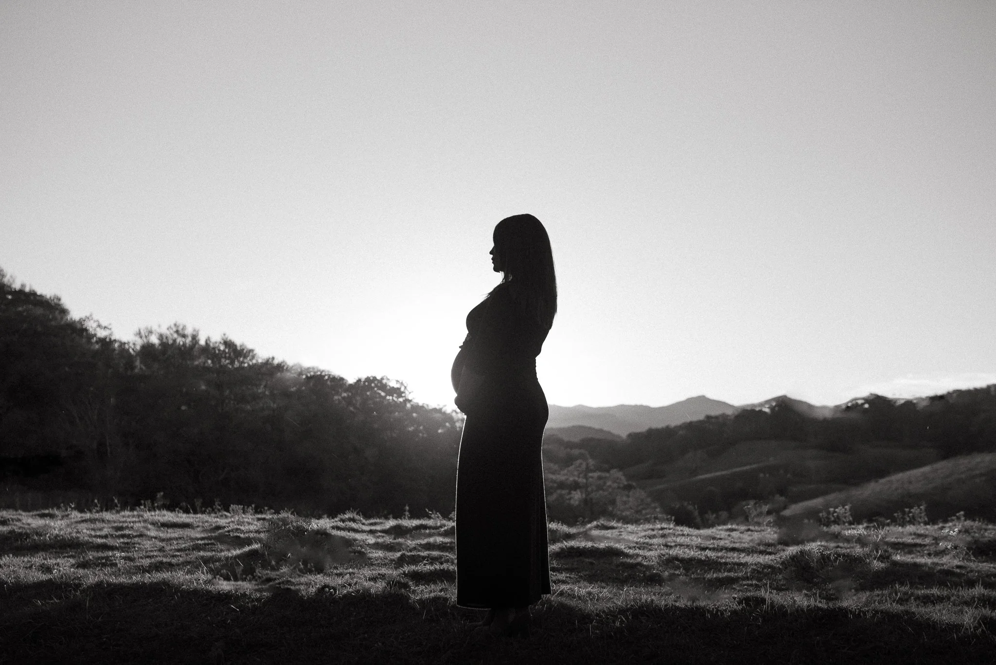 Silhouette of a pregnant woman standing outdoors during sunset or sunrise, with mountains and trees in the background.
