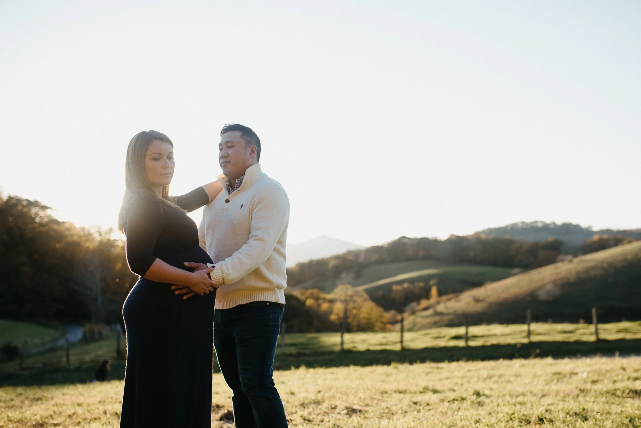 A couple holding each other affectionately in a scenic outdoor landscape with rolling hills and trees during sunset.