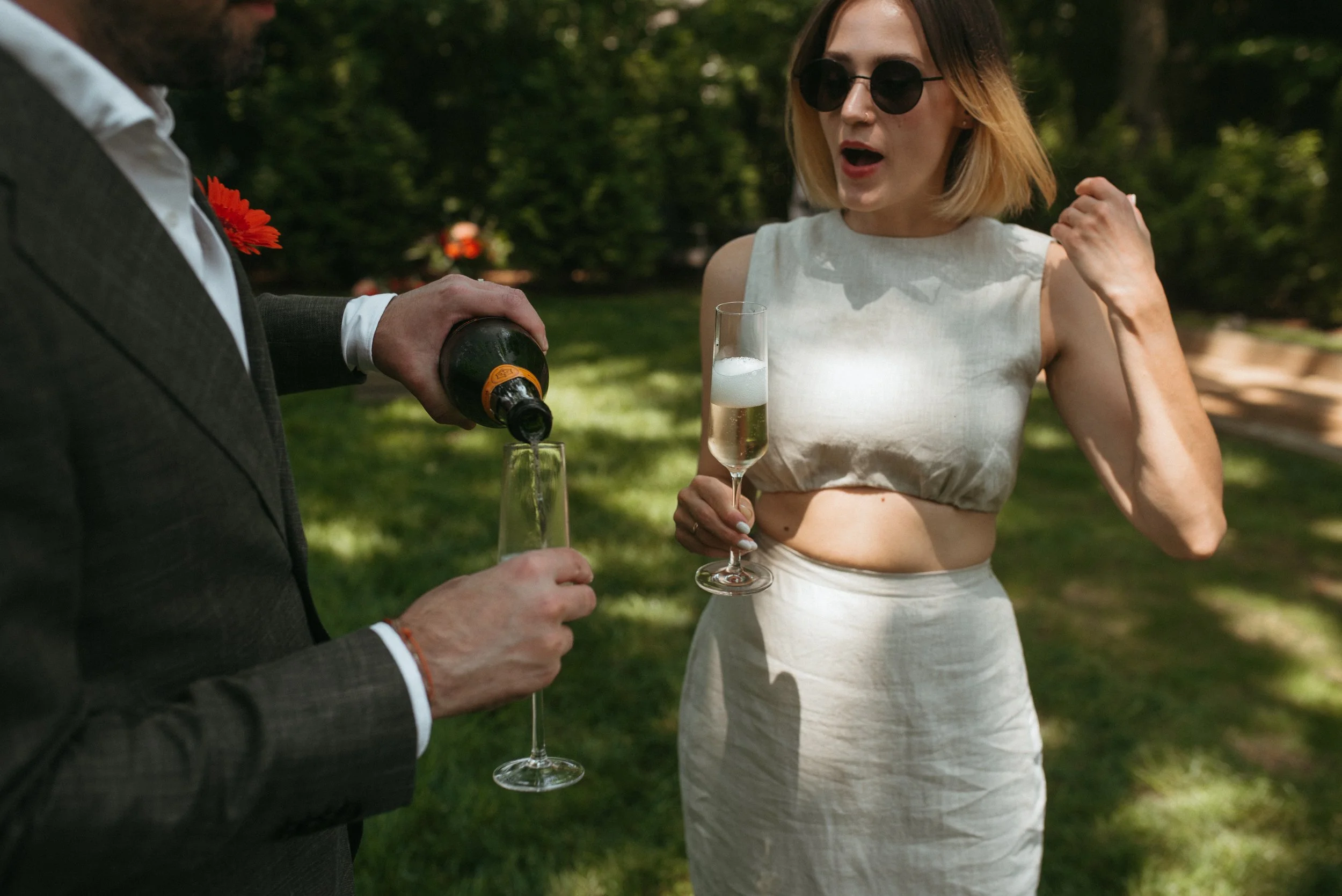 A woman in a white dress and sunglasses holding a champagne flute, receiving a bottle of champagne poured into her glass from a man in a gray suit with a red flower on his lapel, outdoors in a green park.