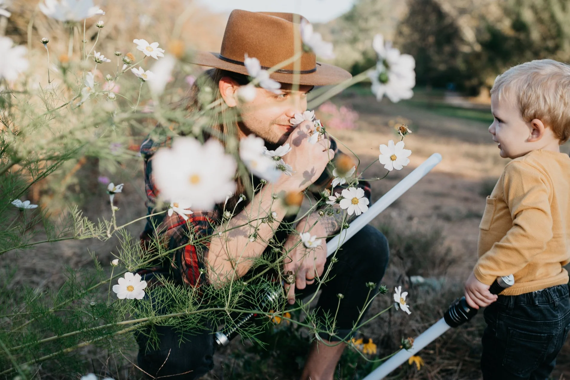 A man with a brown fedora hat and a plaid shirt crouches among white daisy flowers, smelling one close to his nose, while a young boy with blonde hair and a yellow shirt stands nearby holding a white pole with a black handle, engaging with the man outdoors in a garden during daylight.