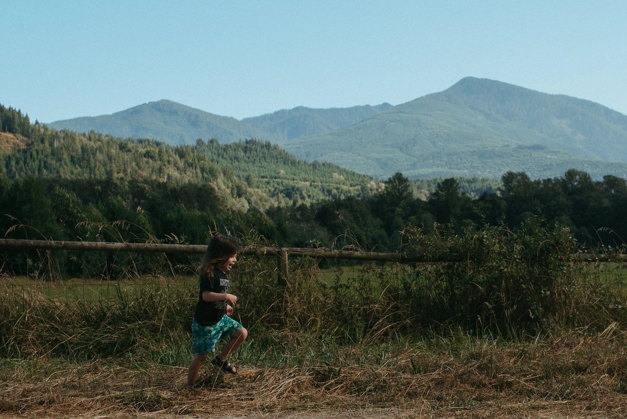 A young boy running through tall grass with mountains in the background under a clear sky.
