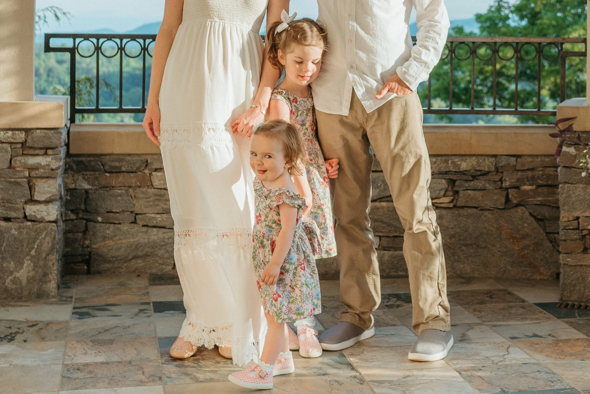 A family of three children standing on a stone patio with a stone railing, with a scenic view of green trees and mountains in the background. The children are wearing casual summer clothes and smiling.
