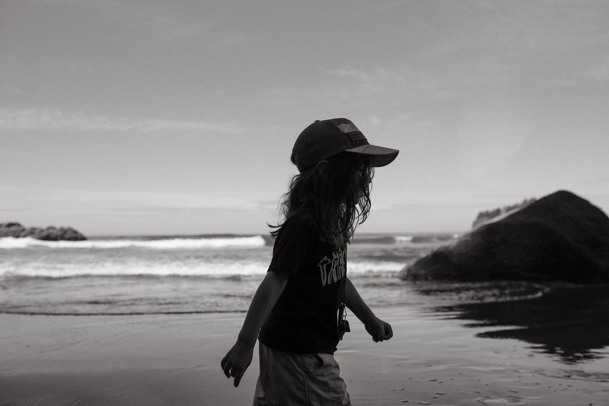 A person wearing a baseball cap and a t-shirt walking along the beach at sunset or sunrise, with waves and rocky formations in the background.