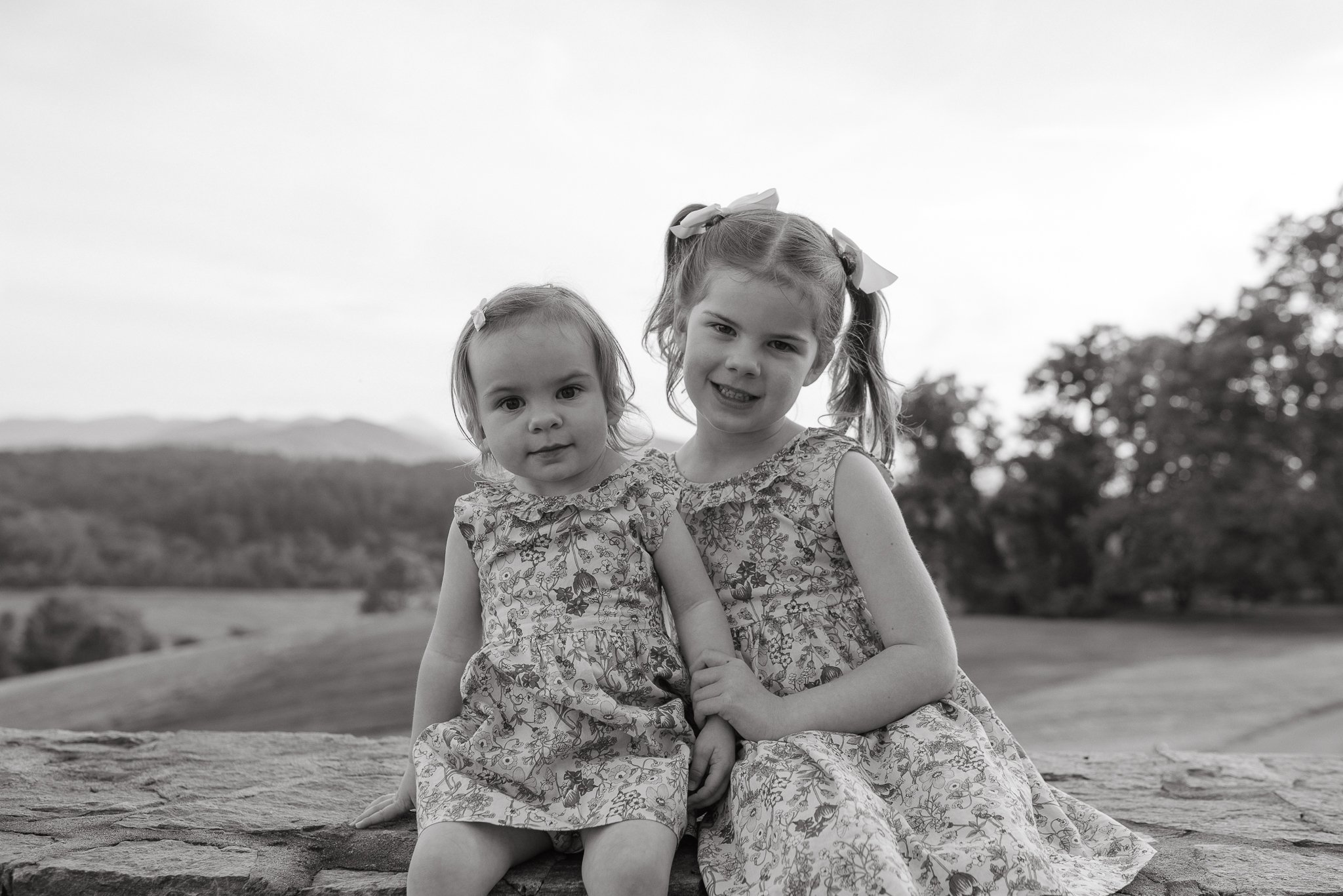Two young girls with light skin, wearing floral dresses with bows in their hair, sitting outdoors on a flat rock with a scenic background of grass, trees, and distant hills, captured in black and white.