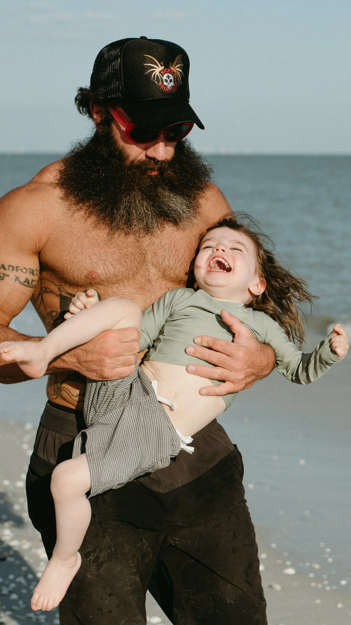 A bearded man wearing a trucker hat and sunglasses holds a laughing young girl at the beach, with the ocean and sky in the background.