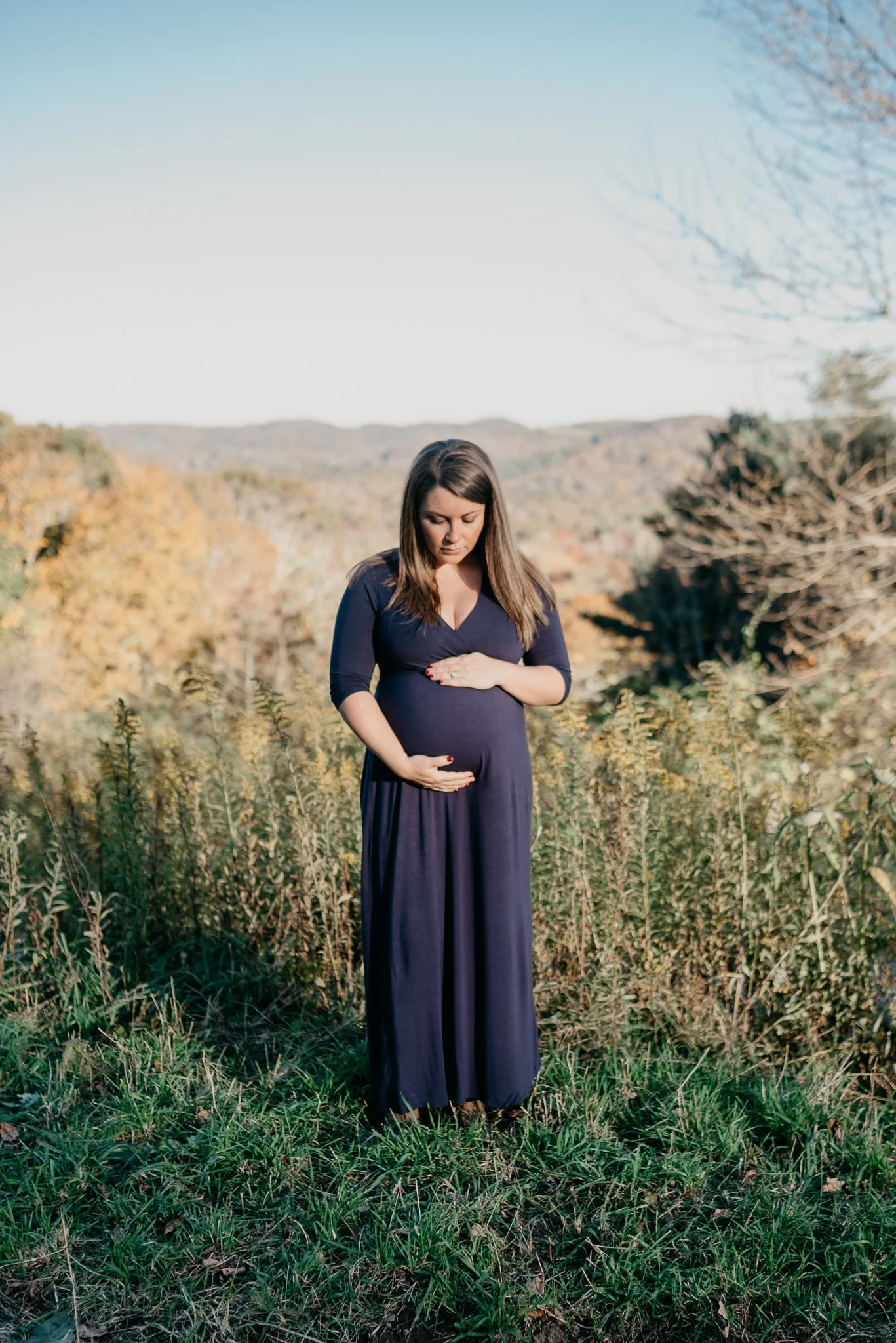 A pregnant woman in a long dark purple dress is standing outdoors in a field with trees and rolling hills in the background. She is holding her belly with both hands and looking down at it.