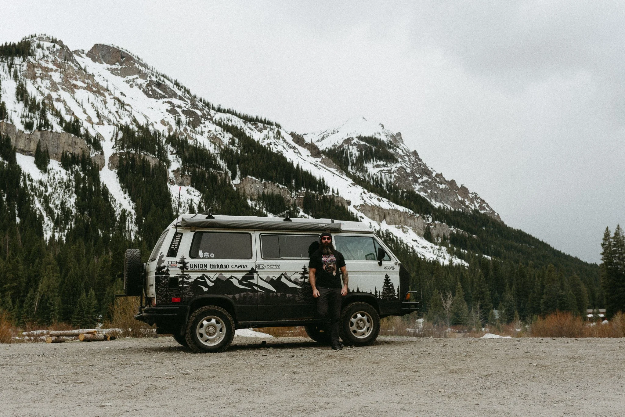 A man with dark hair and beard, wearing sunglasses, a black T-shirt, and dark pants, standing next to a black and white van with mountain and forest graphics, in a mountainous area with snow-covered peaks and evergreen trees.
