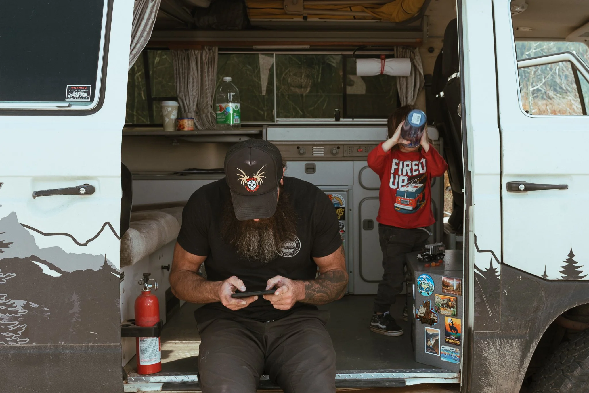 A man with tattoos and a beard wearing a black baseball cap and black shirt sitting inside a camper van, looking at a device. A young boy in a red shirt with a fire truck graphic is standing nearby, drinking from a cup. The camper van interior has cu
