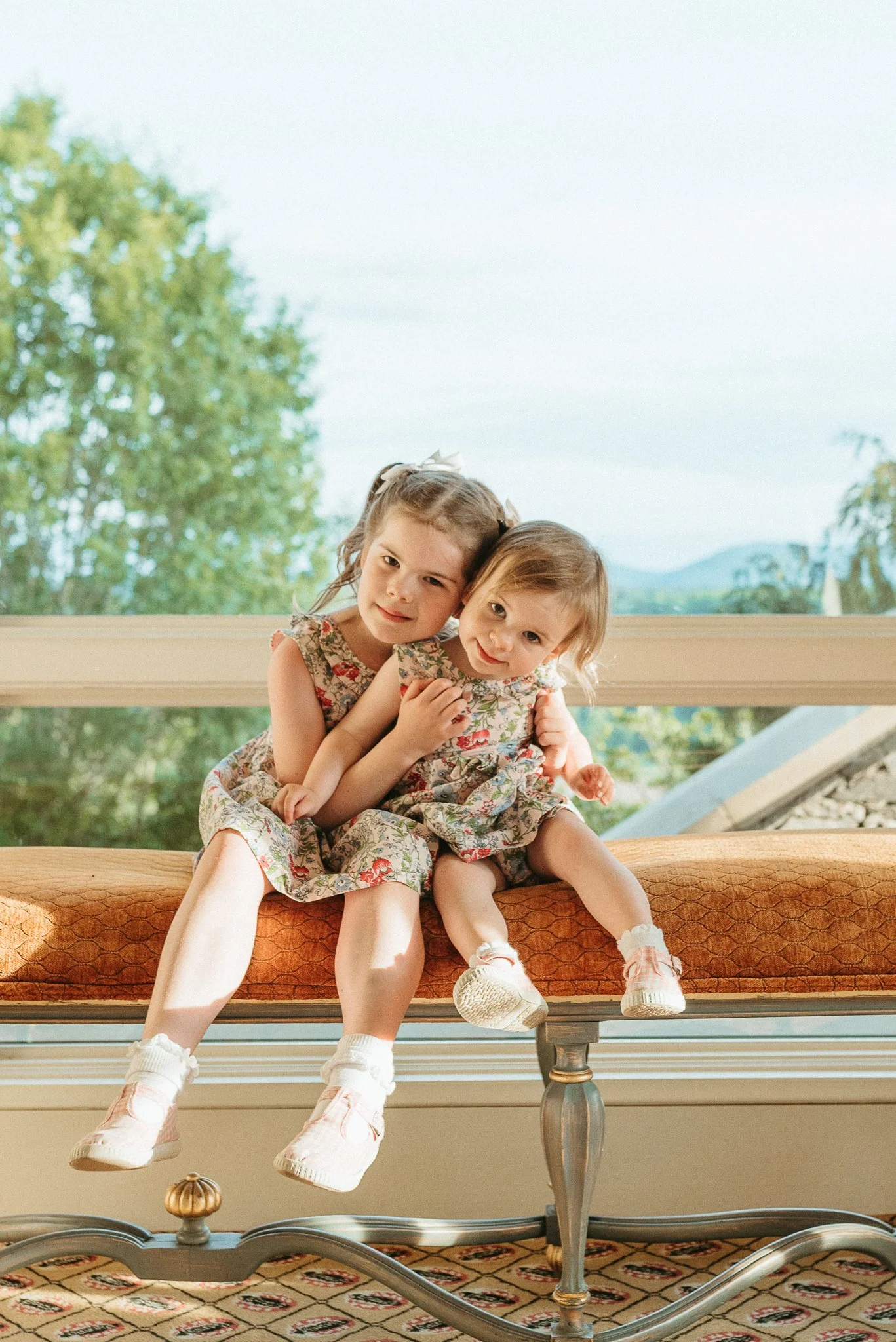 Two young girls in floral dresses sitting on a vintage bench, outdoors with trees and mountains in the background.