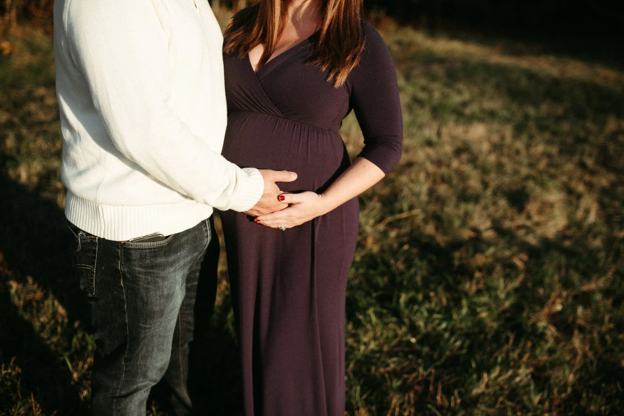 A man and a pregnant woman are standing close together outdoors, with the man's hand gently resting on the woman's belly. Only their torsos and part of their arms are visible.