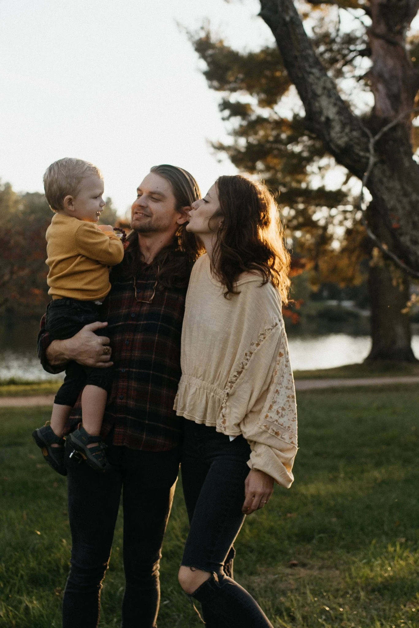 A family of three sharing a special moment outdoors during sunset. The father is holding their young son, who is looking at his parents. The mother stands close, the scene is set near a lake with trees in the background.