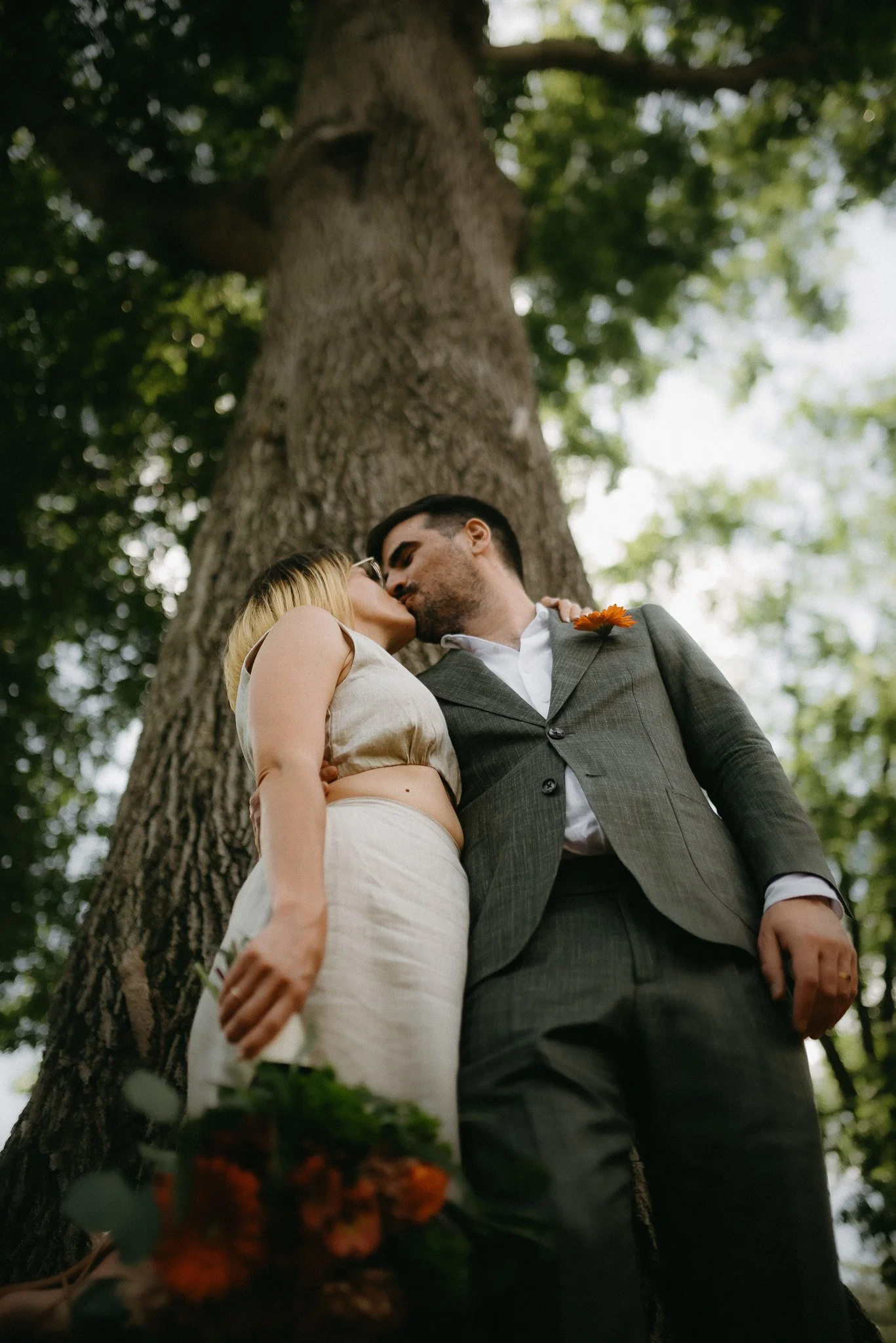 A couple sharing a tender kiss outdoors under a large tree, with the woman wearing glasses and a cream-colored dress, and the man in a gray suit with a boutonniere, surrounded by green foliage.
