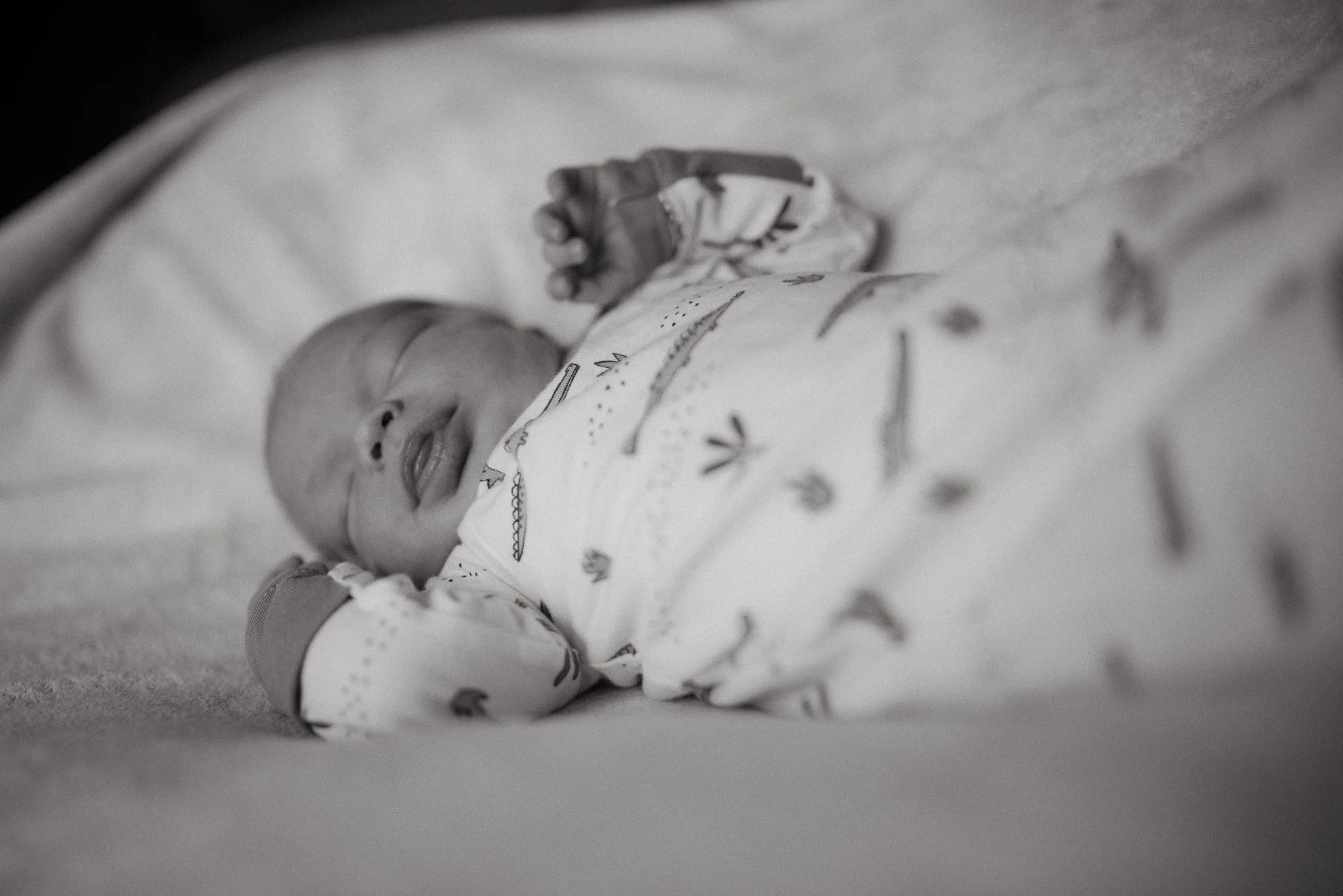 Black and white photo of a newborn baby lying on a blanket, with a hand gently holding the baby’s hand.