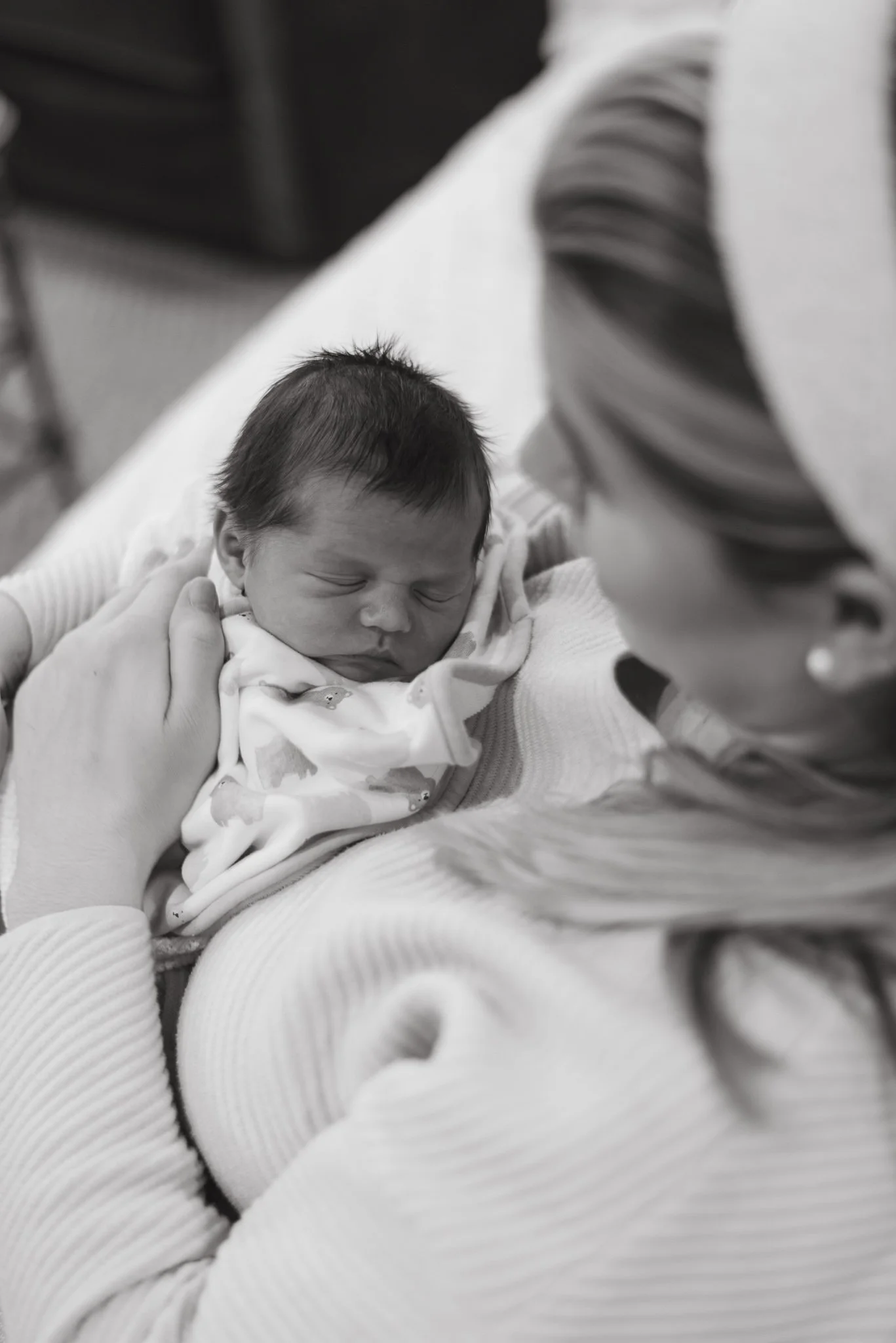 A newborn baby with dark hair sleeping, held by an adult wearing a white sweater, with a person in a striped shirt nearby.