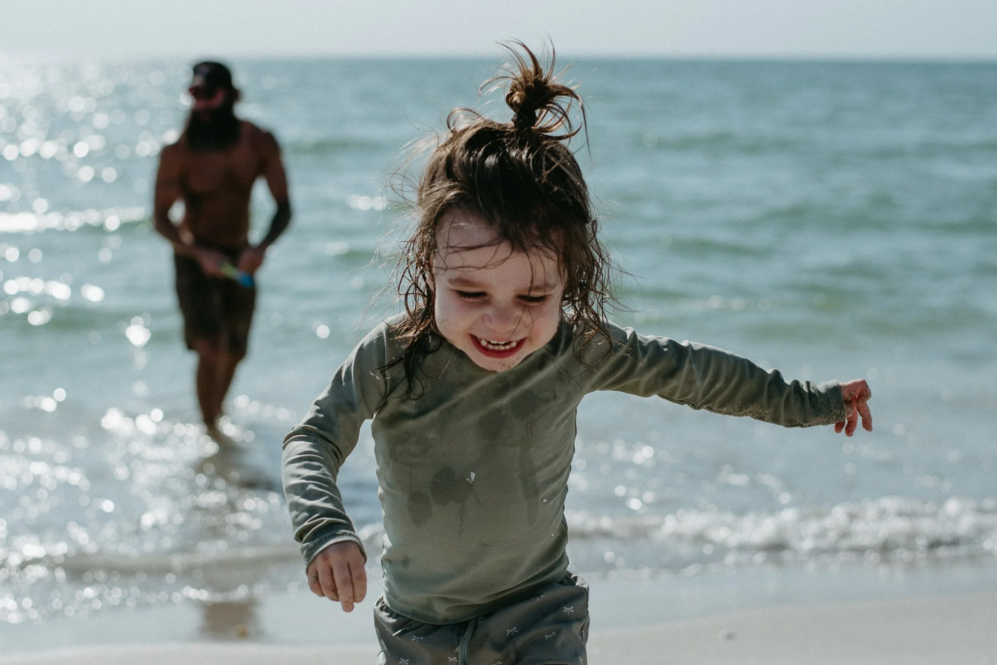 A young girl with wet hair smiling and playing at the beach, with a man in the background near the water.